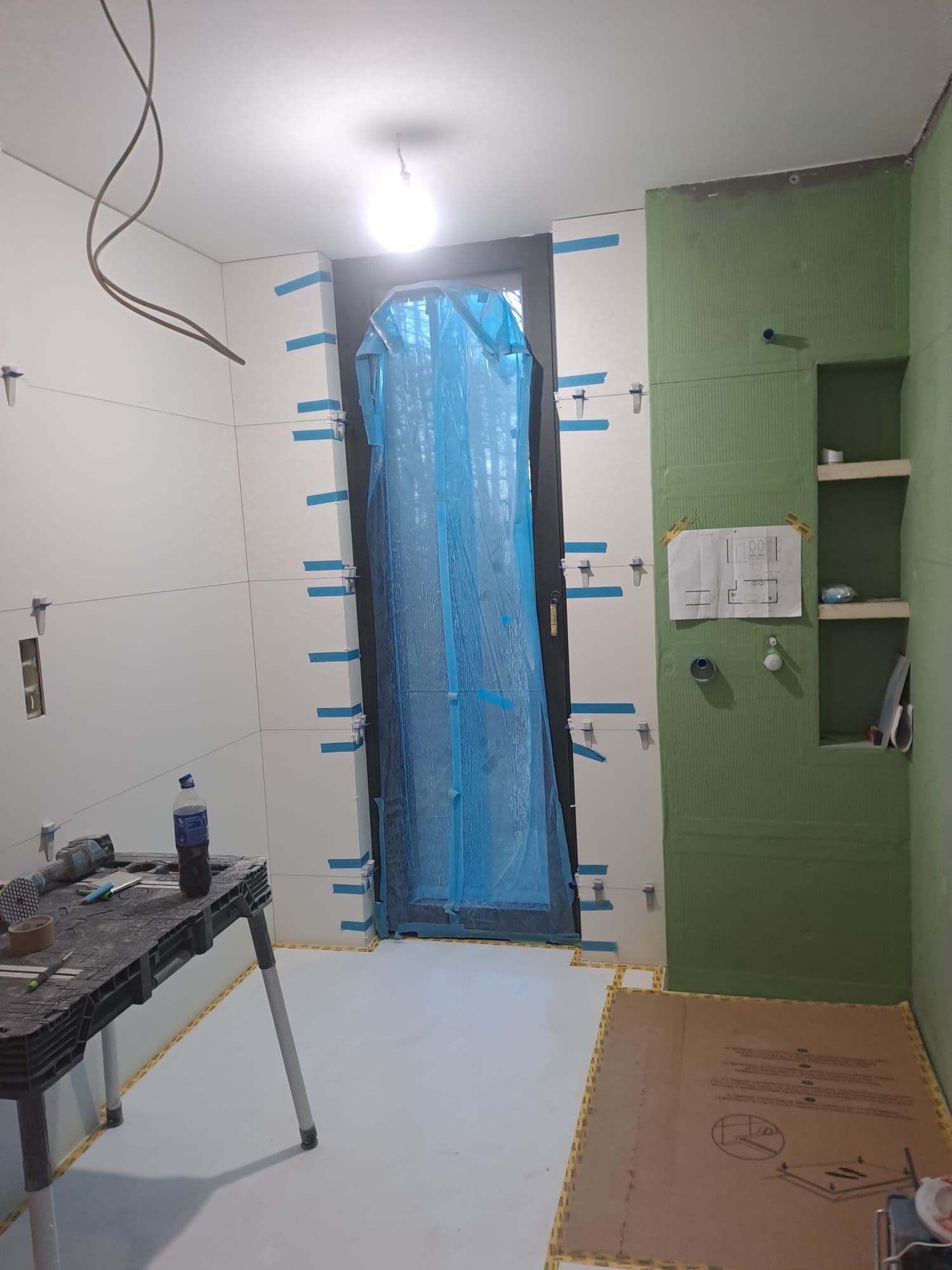 Łazienka w trakcie prac glazurniczych: białe płytki na ścianach, zielona płyta budowlana, narzędzia na stole. Drzwi zabezpieczone folią. Widoczny plan łazienki.