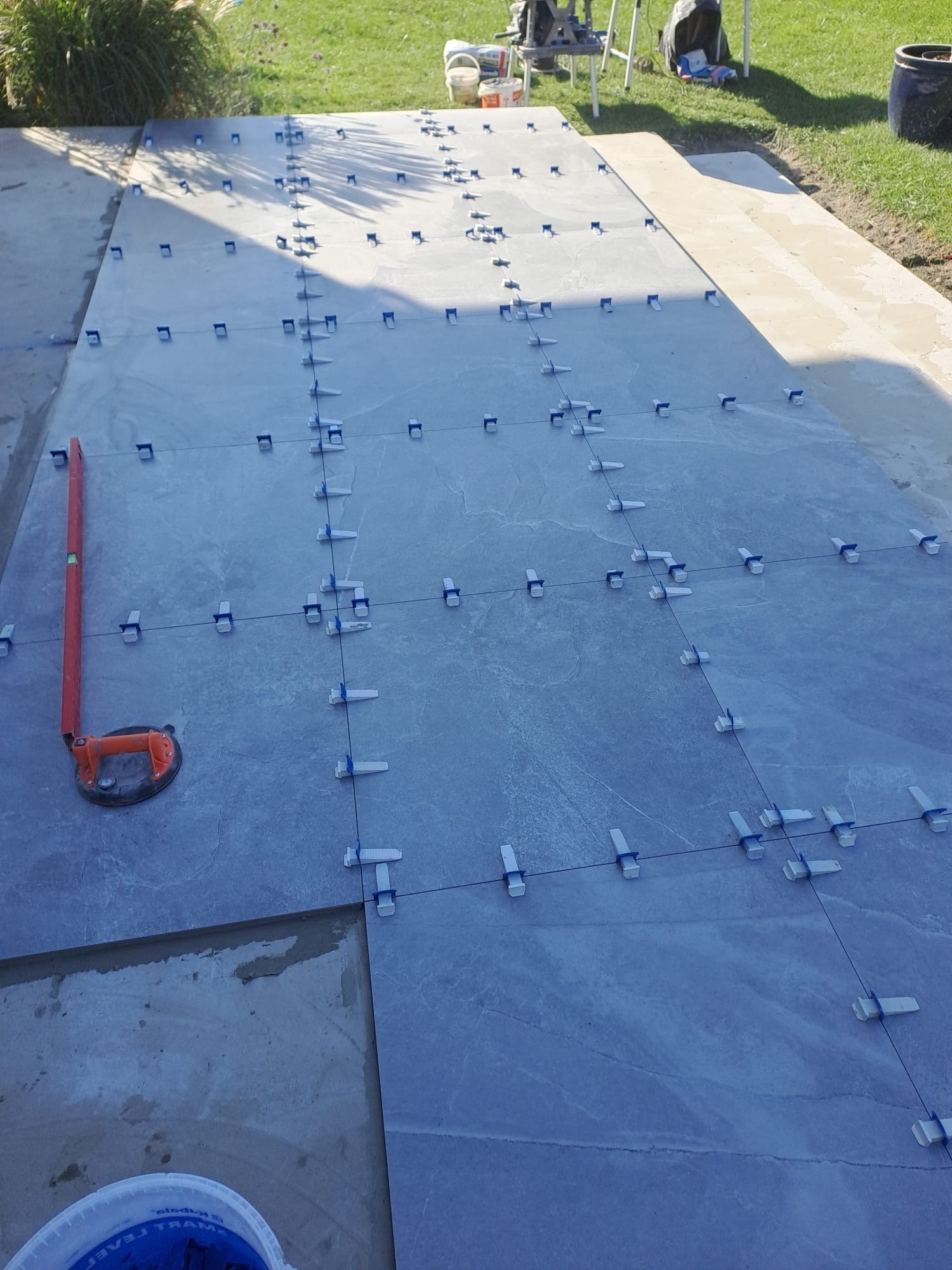 Układanie szarych płytek na zewnątrz z użyciem poziomicy i klipsów poziomujących, widoczny fragment niezakończonej powierzchni.