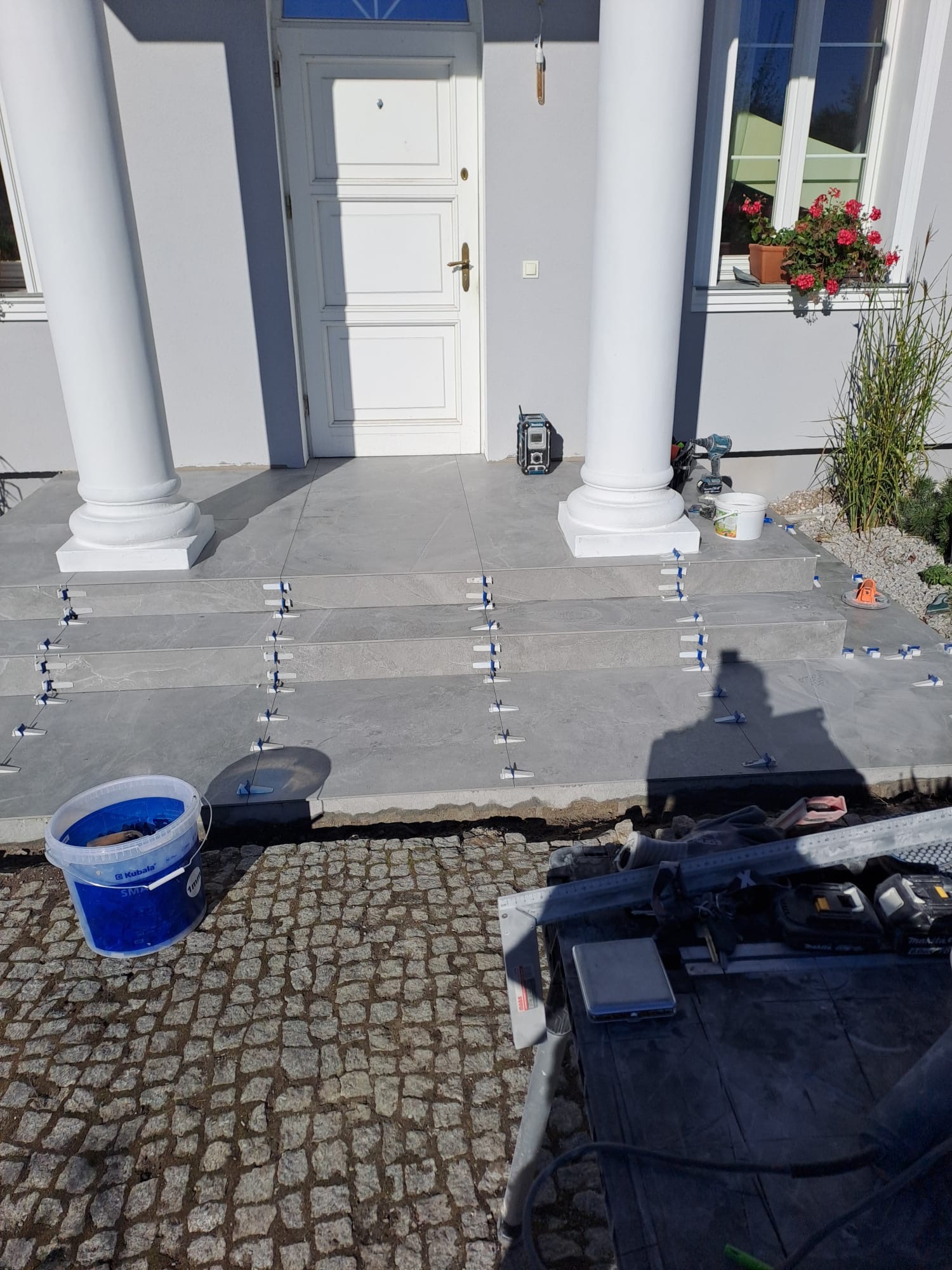 Układanie płytek na schodach wejściowych do domu z kolumnami. Widoczne narzędzia glazurnicze i system poziomowania płytek. Praca w toku, etap wykończeniowy.