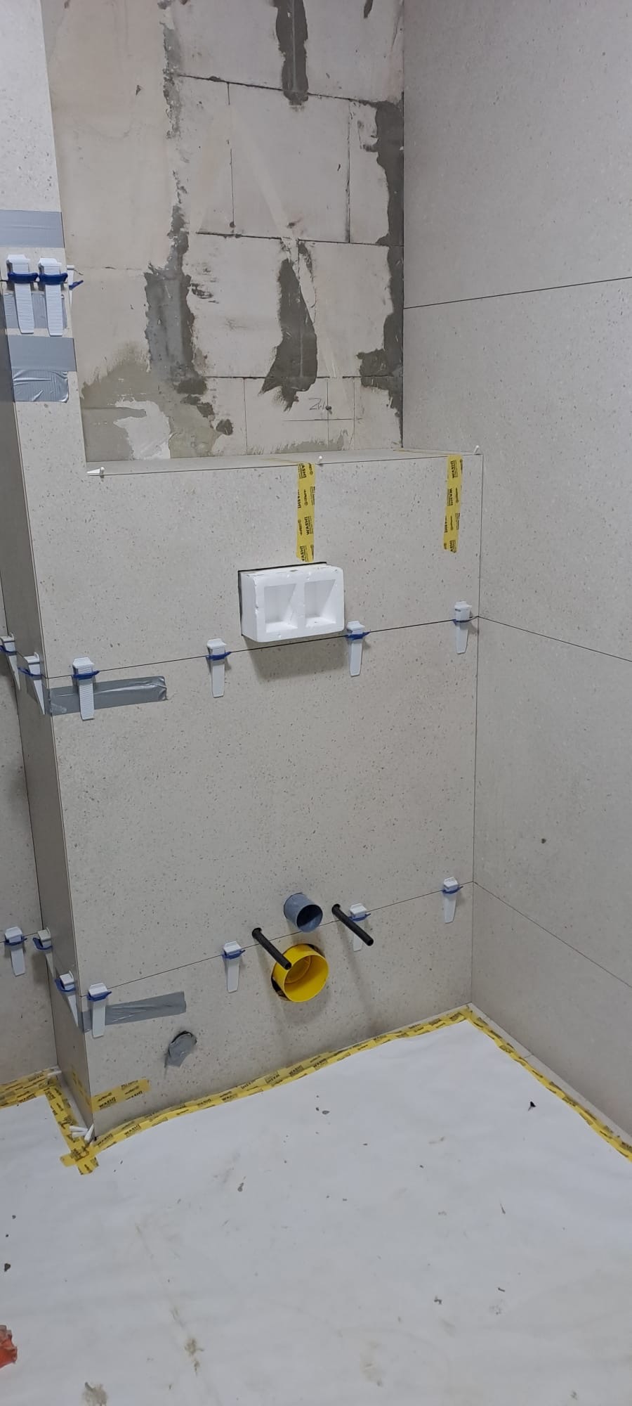 Fragment łazienki w trakcie prac glazurniczych: ułożone płytki na ścianach, widoczne elementy instalacji wodnej i poziomice, podłoga zabezpieczona folią.