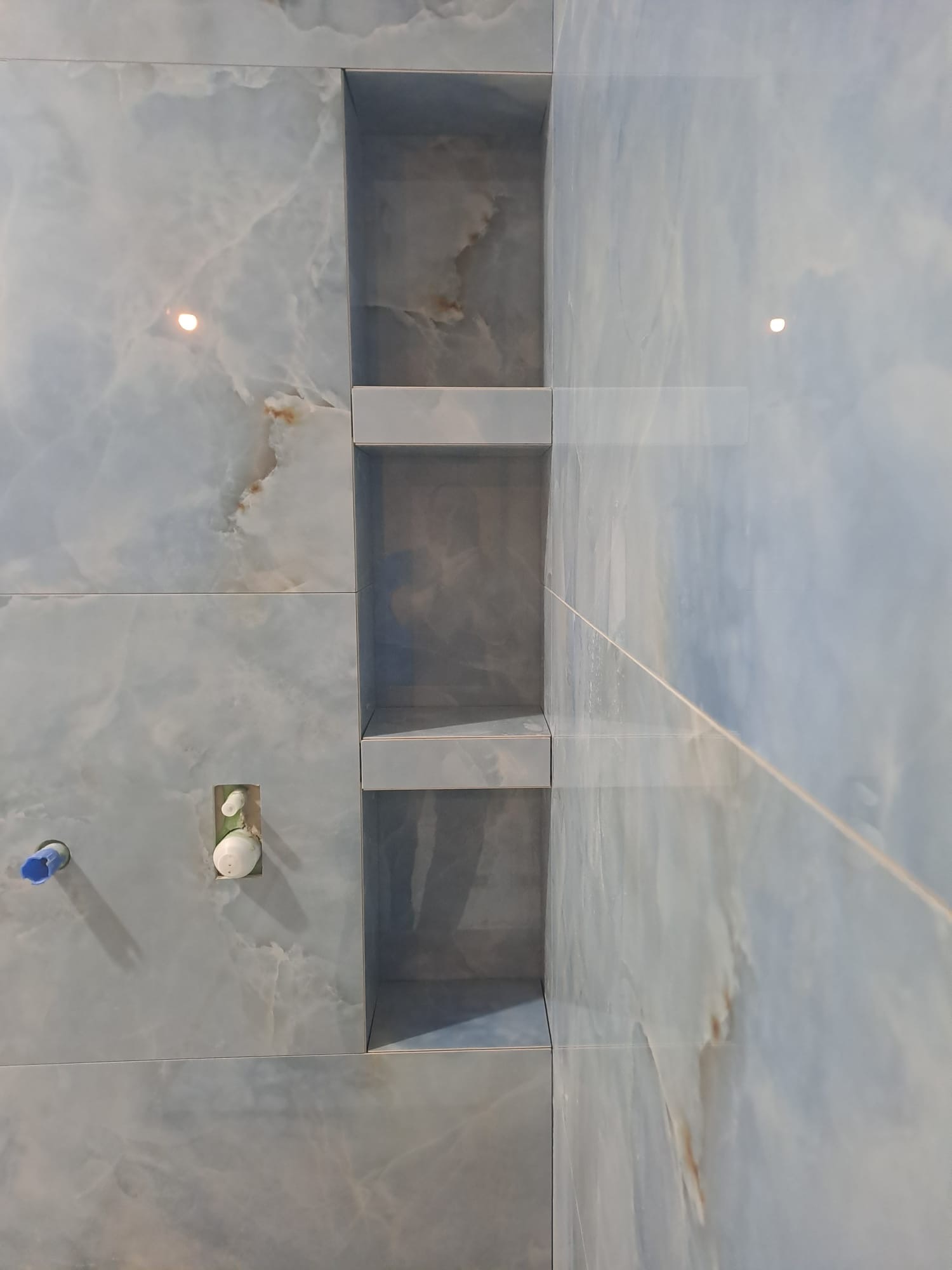 Niebieskie płytki imitujące marmur z wnęką prysznicową z półkami. Widoczne przyłącza wodne. Nowoczesne wykończenie łazienki z dbałością o detale.