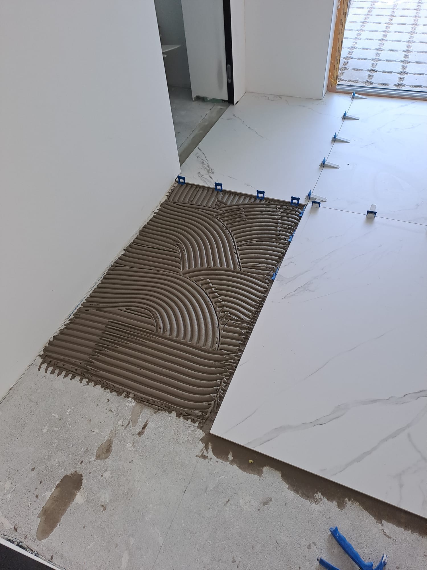 Układanie płytek podłogowych z użyciem systemu poziomowania w Szczecinie. Widoczna nałożona zaprawa i płytki imitujące marmur, w tle drzwi i okno.