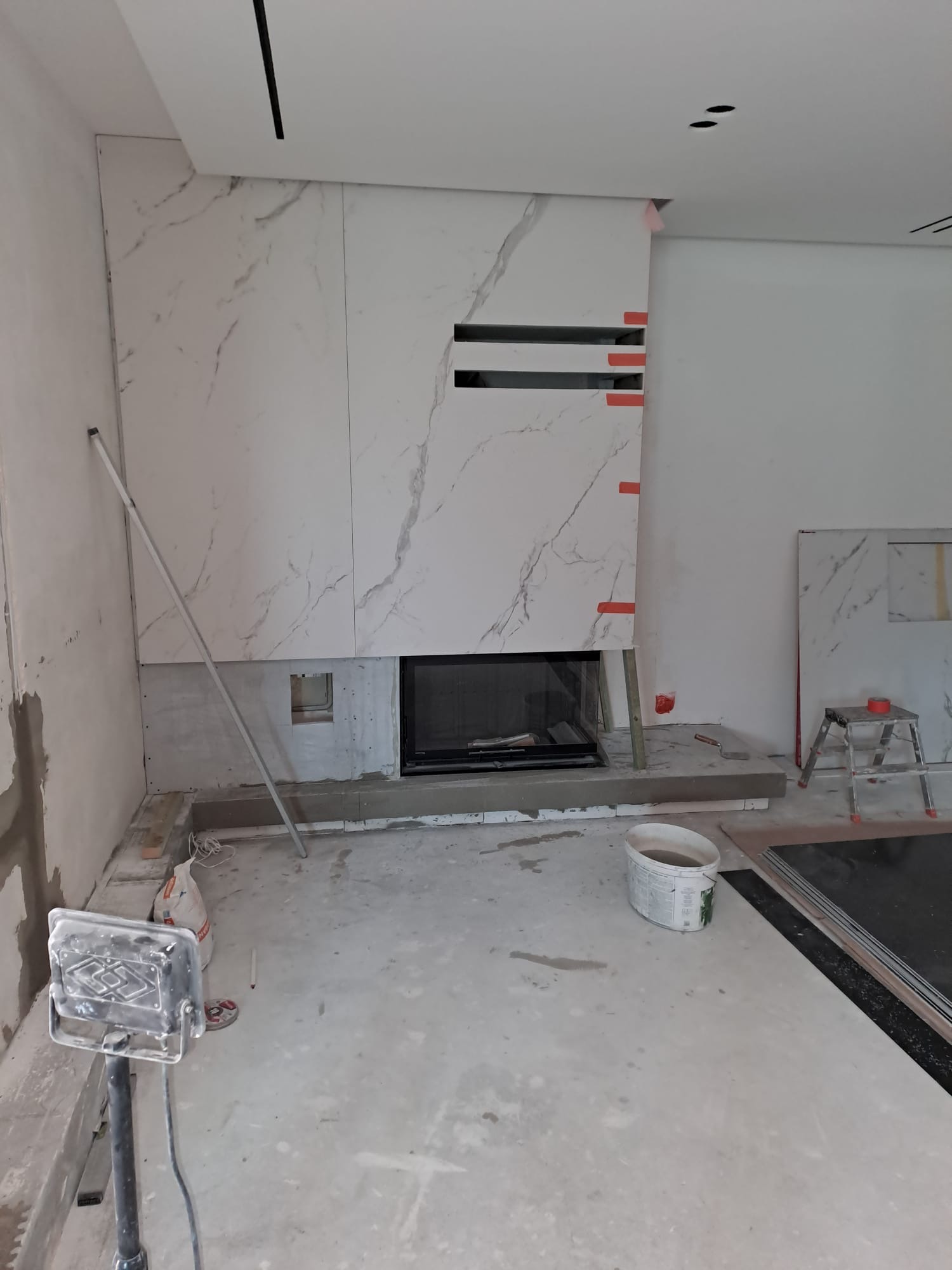 Montaż okładziny imitującej marmur na ścianie z kominkiem. Widoczne narzędzia i materiały budowlane. W tle fragment ściany i sufitu z oświetleniem.