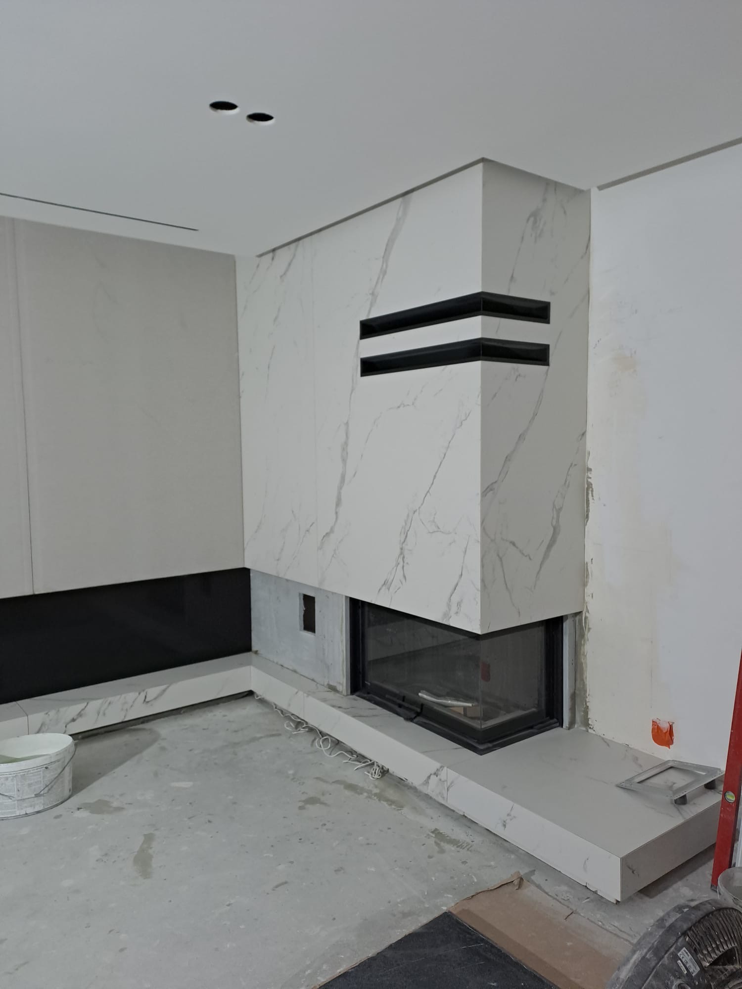 Nowoczesny kominek obłożony białym marmurem z czarnymi akcentami, w trakcie wykończenia, widoczne narzędzia i wiadro z farbą. Element wystroju wnętrza.