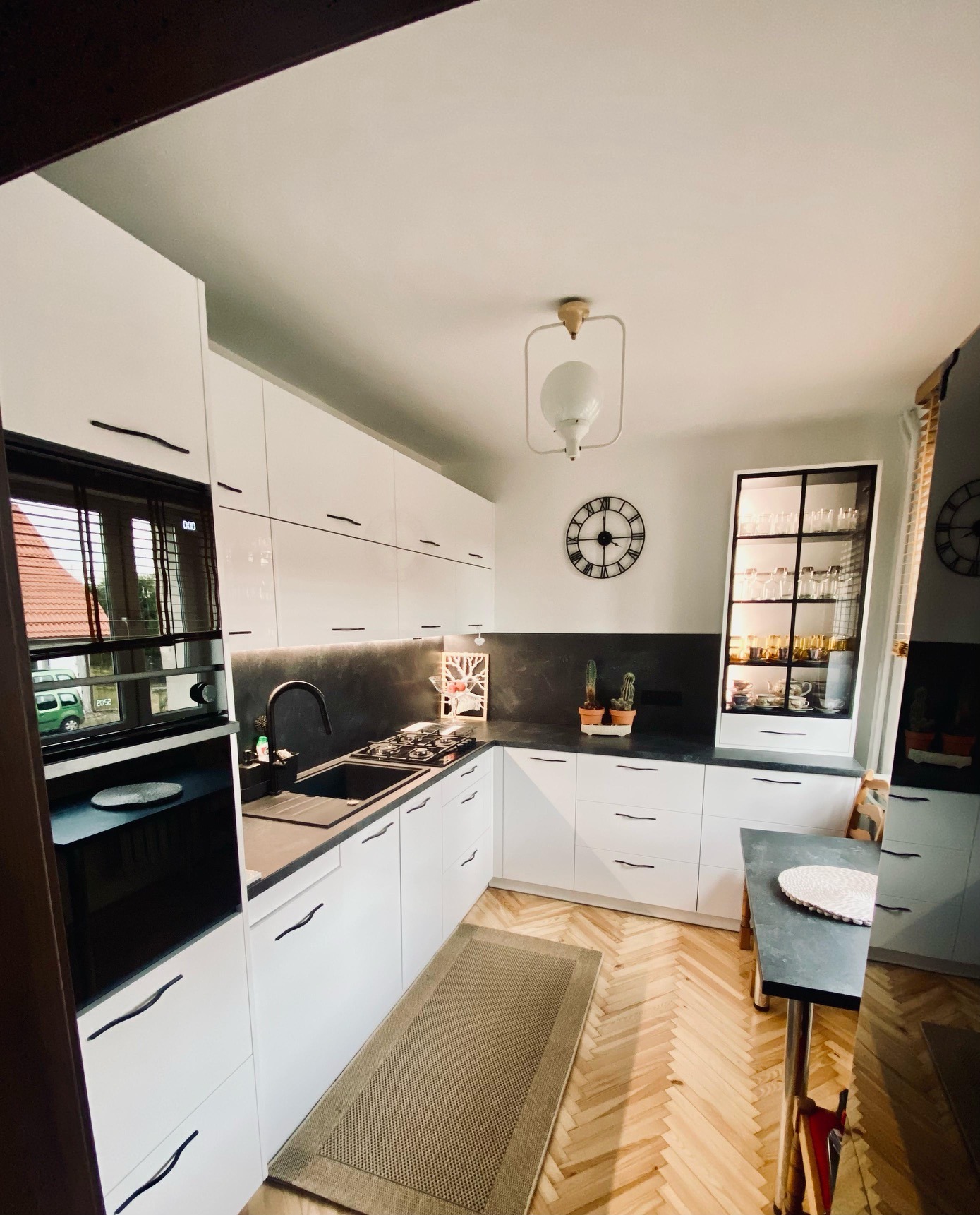 Nowoczesna kuchnia z białymi frontami i czarnym blatem w Szczecinku. Widoczny piekarnik, płyta grzewcza i szafka z przeszklonym frontem. Parkiet w jodełkę.