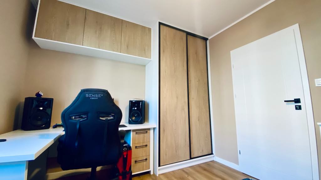 Szafa wnękowa z przesuwnymi drzwiami w pokoju gracza, zintegrowana z biurkiem i szafkami wiszącymi w odcieniach drewna i bieli. Widoczny fotel gamingowy.