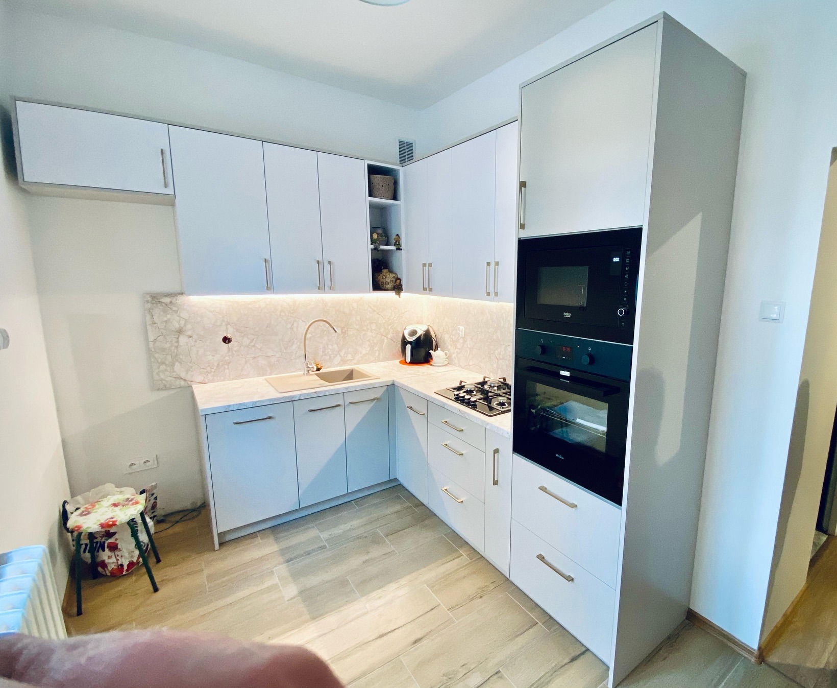 Nowoczesna, jasna kuchnia z białymi szafkami, marmurowym blatem i oświetleniem LED. Widoczna płyta gazowa, piekarnik i mikrofalówka w zabudowie. Minimalistyczny design.