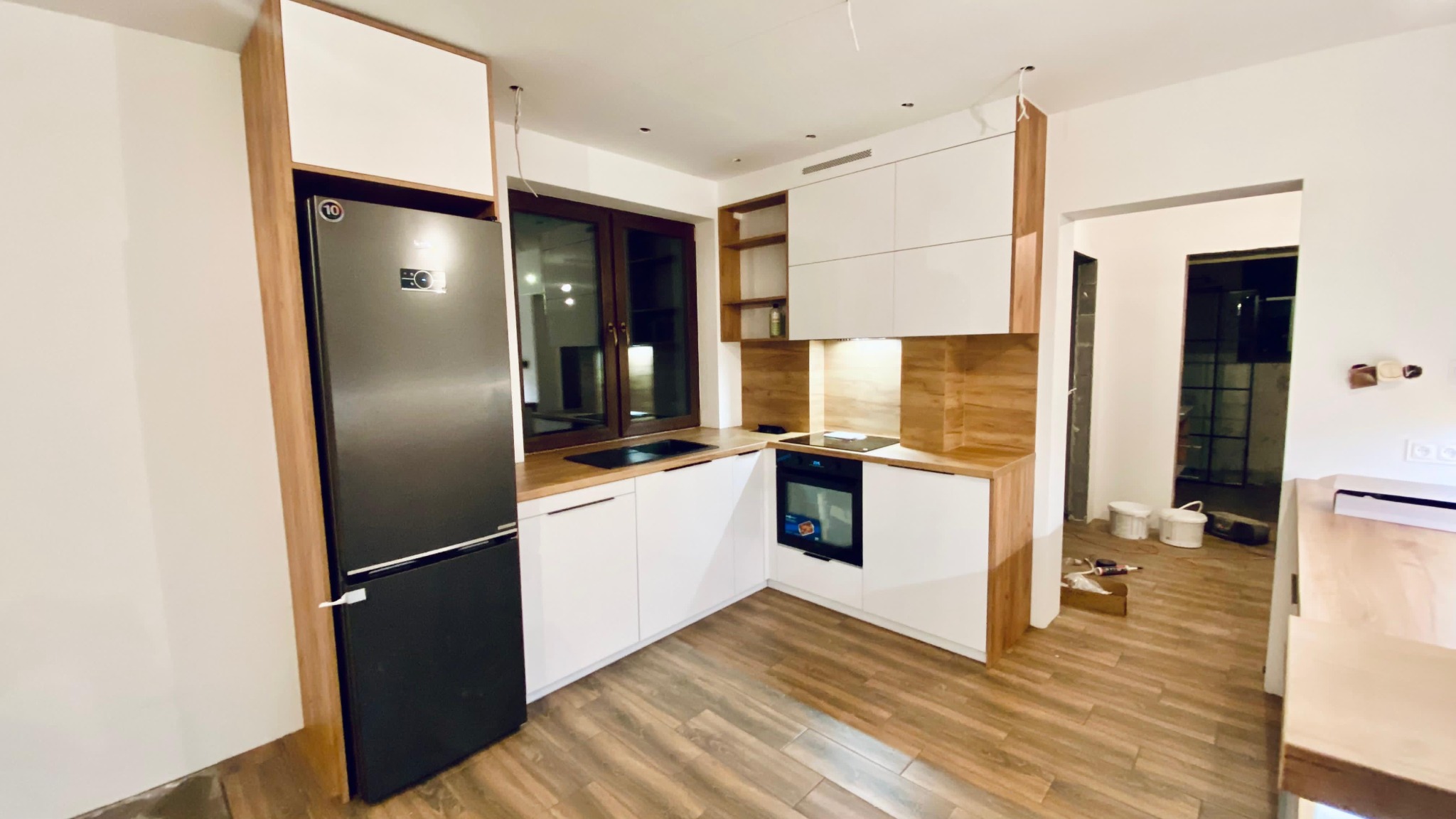 Nowoczesna kuchnia z białymi szafkami i drewnianymi akcentami, czarną lodówką i blatem z drewnopodobnego laminatu, widok na okno i fragment podłogi.