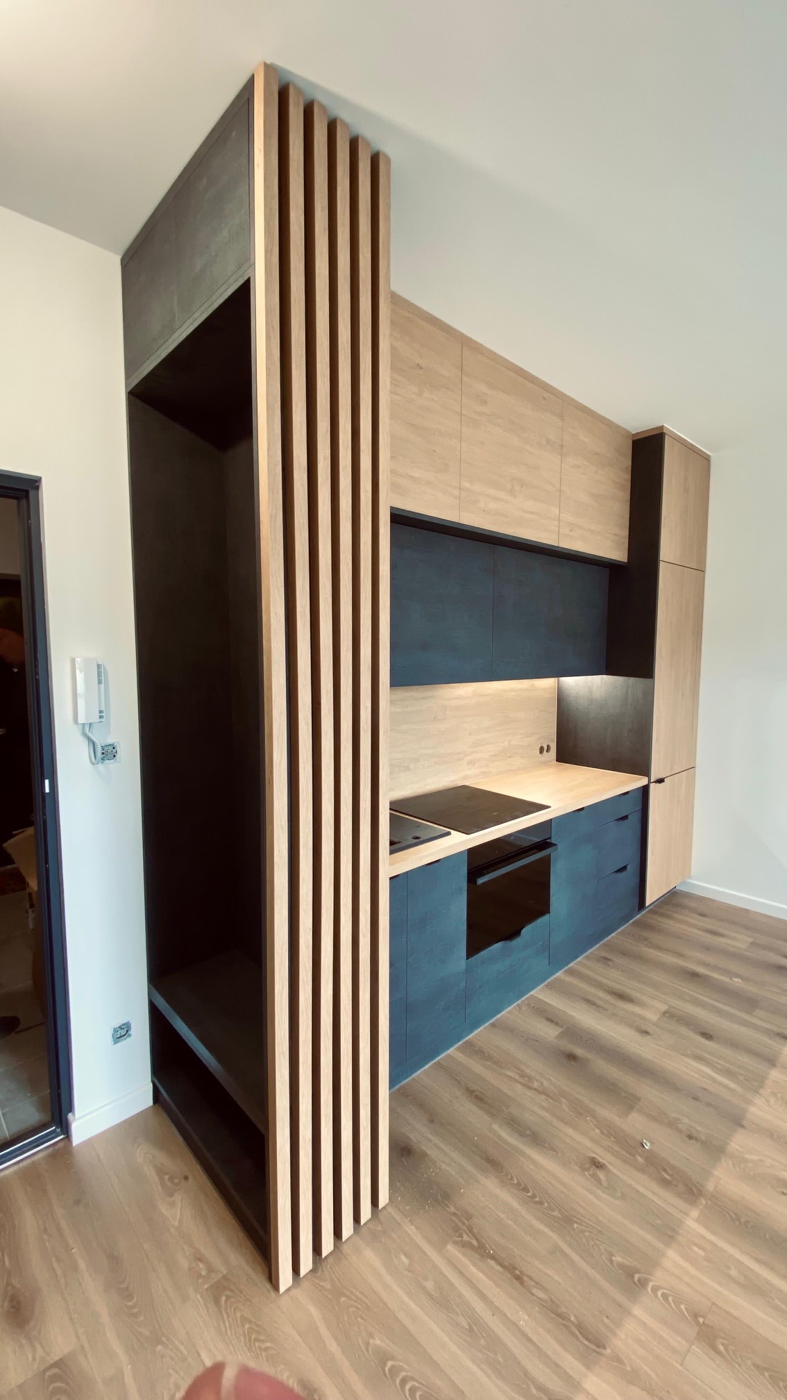 Nowoczesna kuchnia z drewnianymi lamelami oddzielającymi otwartą szafkę. Blat z płyty imitującej drewno, dolne szafki w kolorze grafitowym, górne w kolorze jasnego drewna.