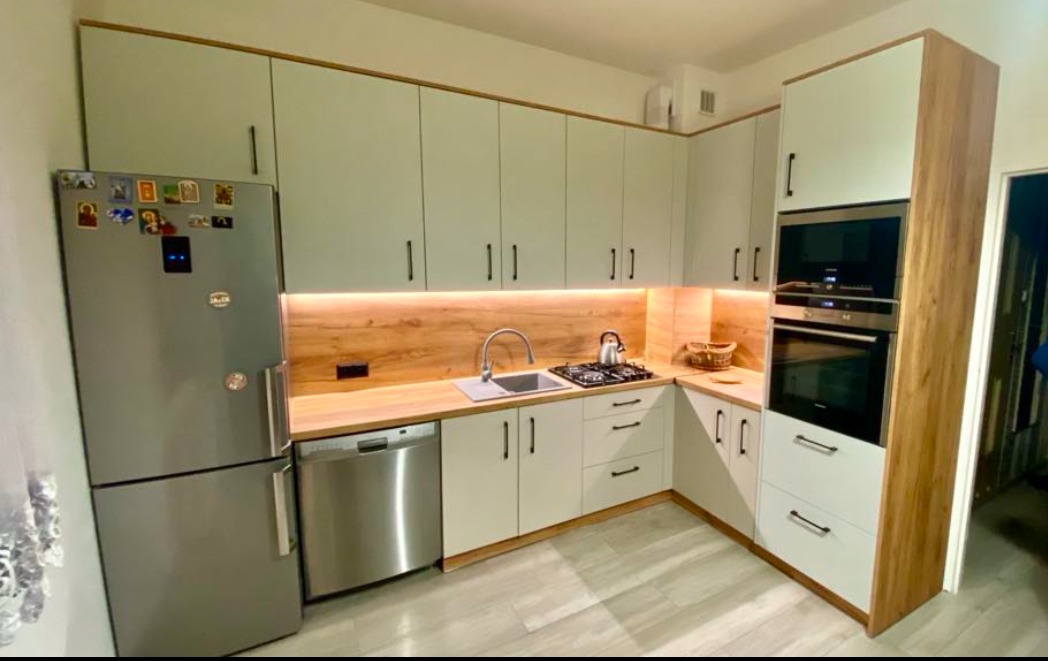 Jasna kuchnia narożna z drewnianym blatem i oświetleniem LED pod szafkami, widoczny srebrny sprzęt AGD i minimalistyczne uchwyty.