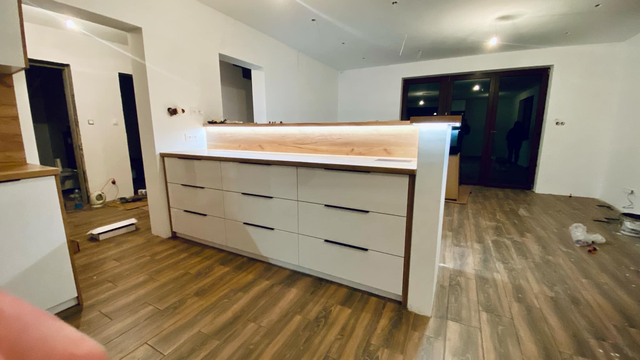 Wyspa kuchenna z białymi frontami i drewnianym blatem, podświetlona LED, na tle wnętrza w trakcie remontu, z widocznymi elementami wykończeniowymi na podłodze.