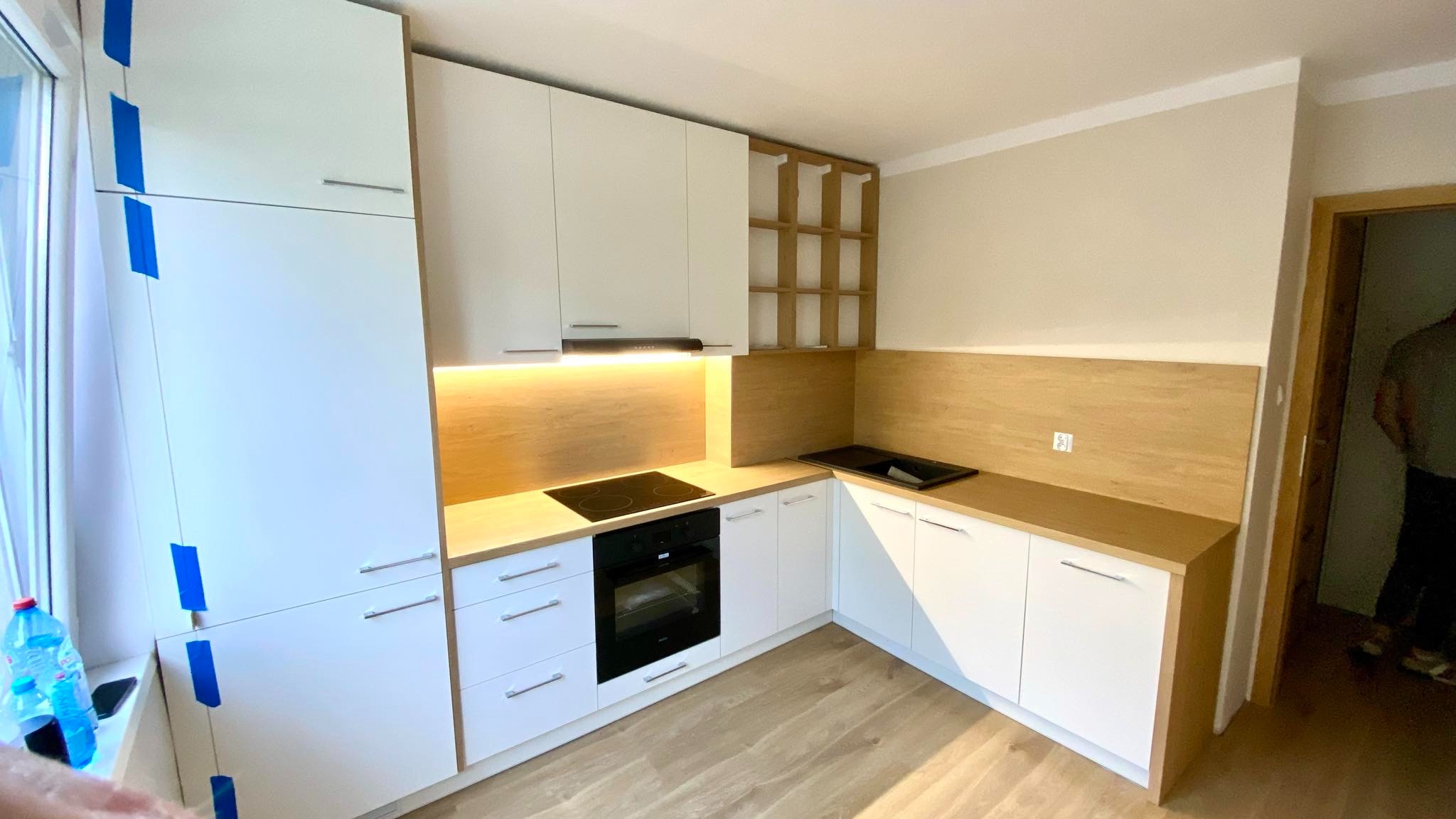 Jasna kuchnia z białymi szafkami i drewnianym blatem, oświetleniem LED pod szafkami. Widoczna płyta indukcyjna i piekarnik, oraz drewniany regał na wino.