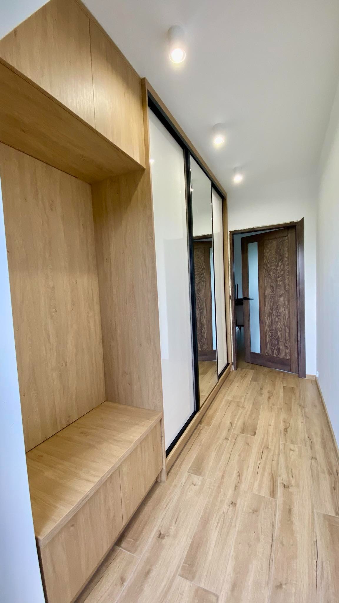 Korytarz z zabudową na wymiar: szafa przesuwna z lustrami i siedzisko z szafkami w odcieniach drewna. Całość dopełnia podłoga imitująca drewno.