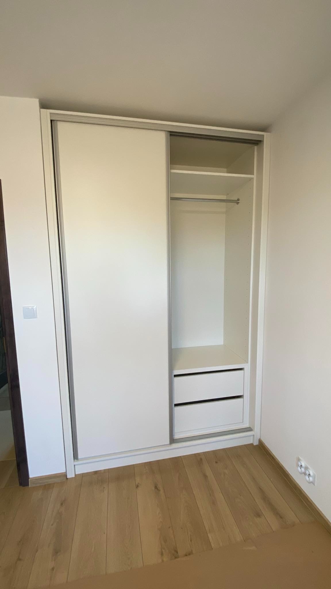 Biała szafa wnękowa z przesuwnymi drzwiami, widoczne dwie szuflady i półka z drążkiem na ubrania. Jasne panele podłogowe. Neutralne tło ścian.
