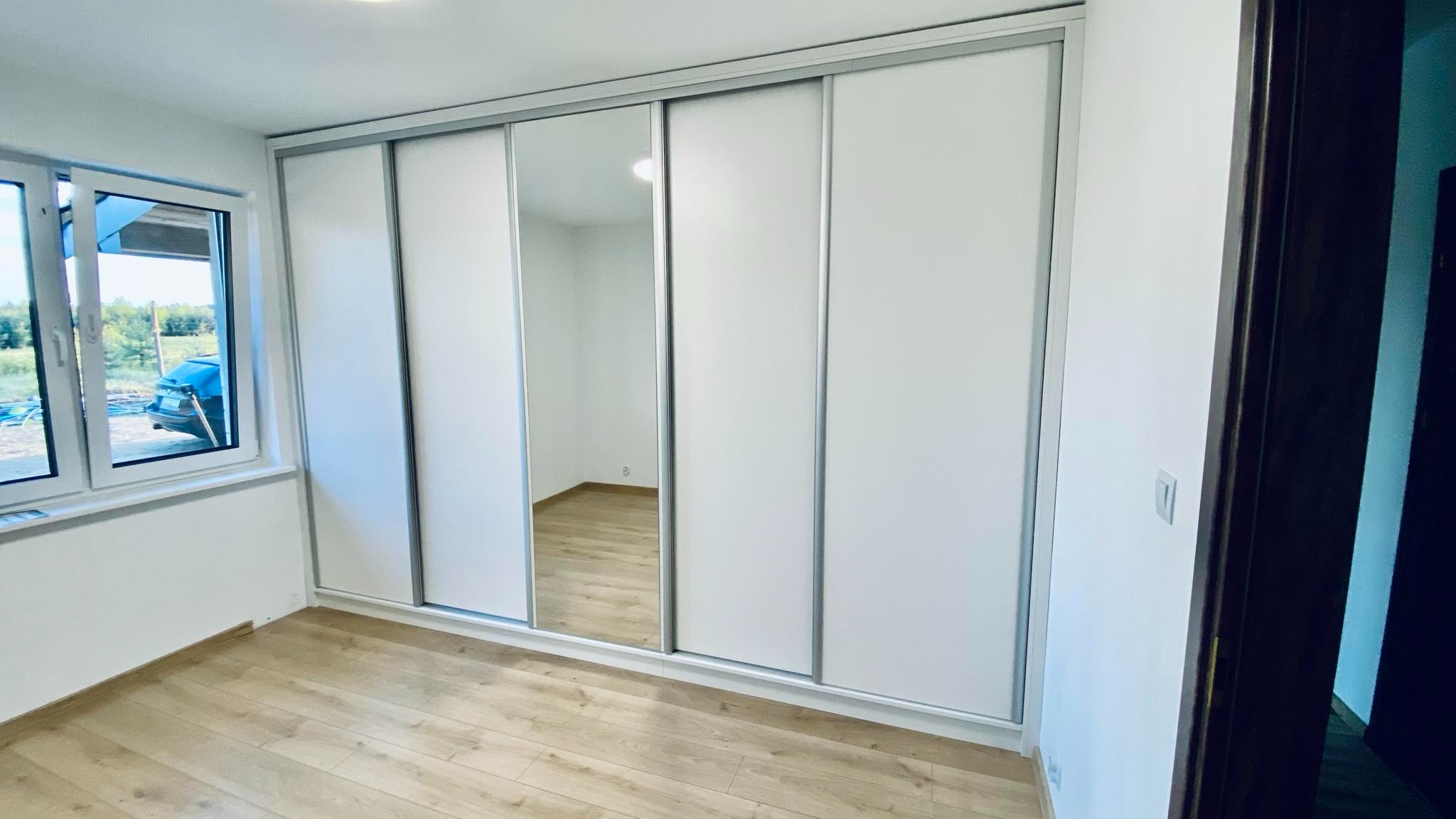 Biała szafa wnękowa z przesuwnymi drzwiami i lustrem, widok z boku, na podłodze panele. W oknie widoczny krajobraz. Minimalistyczny design.