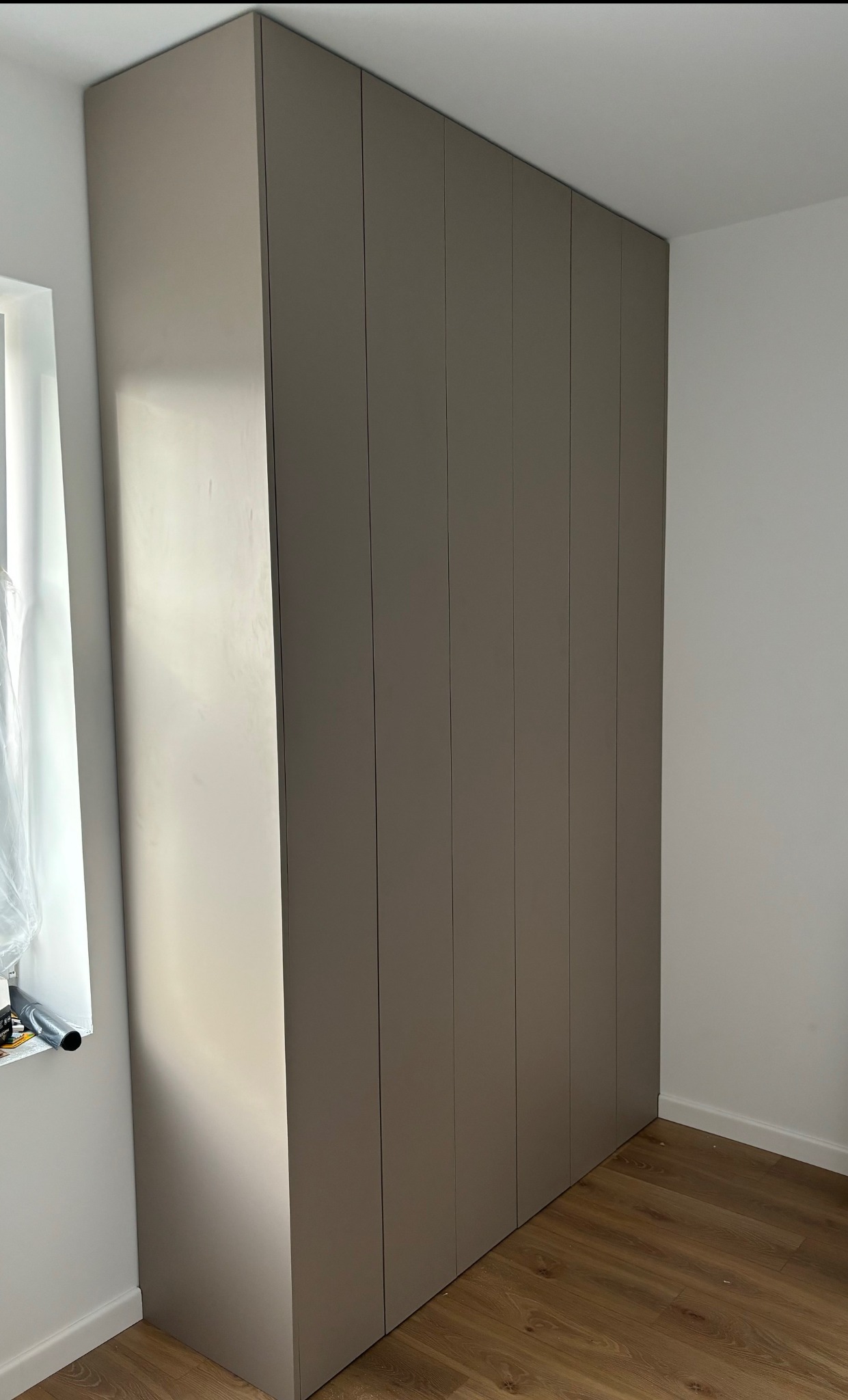 Zamknięta, wysoka szafa wnękowa z szarymi frontami, dopasowana do wnętrza z drewnianą podłogą. Minimalistyczny design i proste linie.