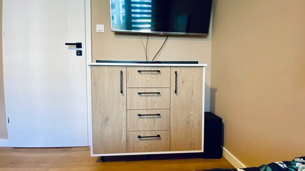 Drewniana komoda z czarnymi uchwytami i białym obramowaniem pod telewizorem, obok białe drzwi z czarną klamką, na tle beżowej ściany.