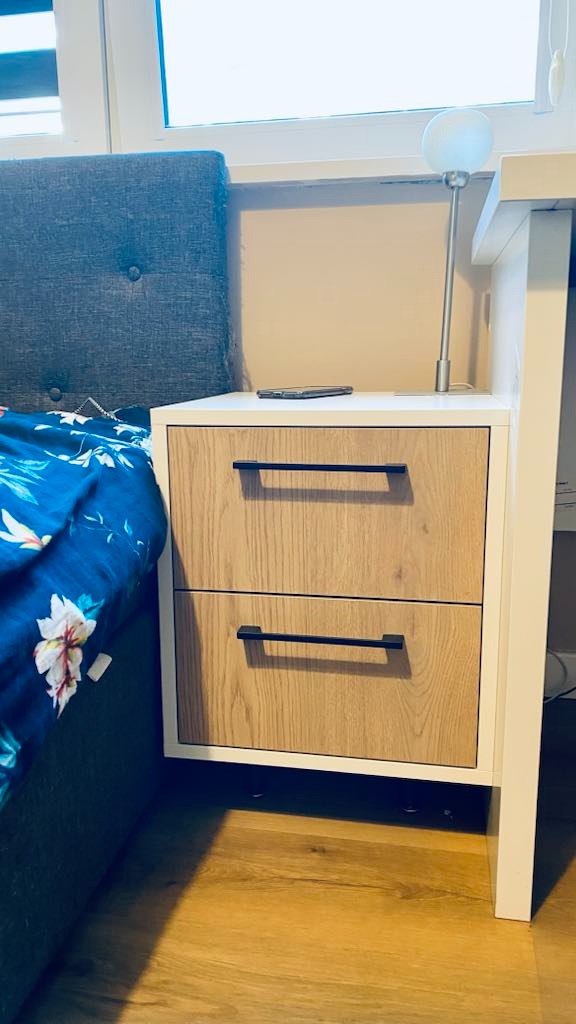 Nowoczesna szafka nocna z dwoma szufladami o drewnianym wykończeniu i czarnymi uchwytami. Na blacie leży telefon, obok lampka. Widoczny fragment łóżka z niebieską pościelą.