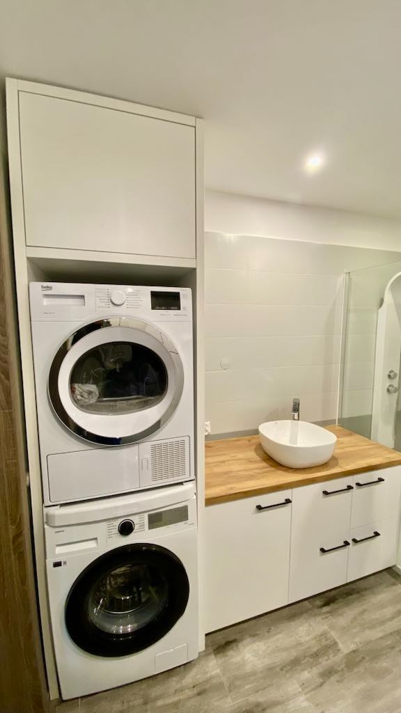 Biała zabudowa łazienkowa z pralką i suszarką w kolumnie, umywalką nablatową i szafkami z drewnianym blatem, minimalistyczny styl.