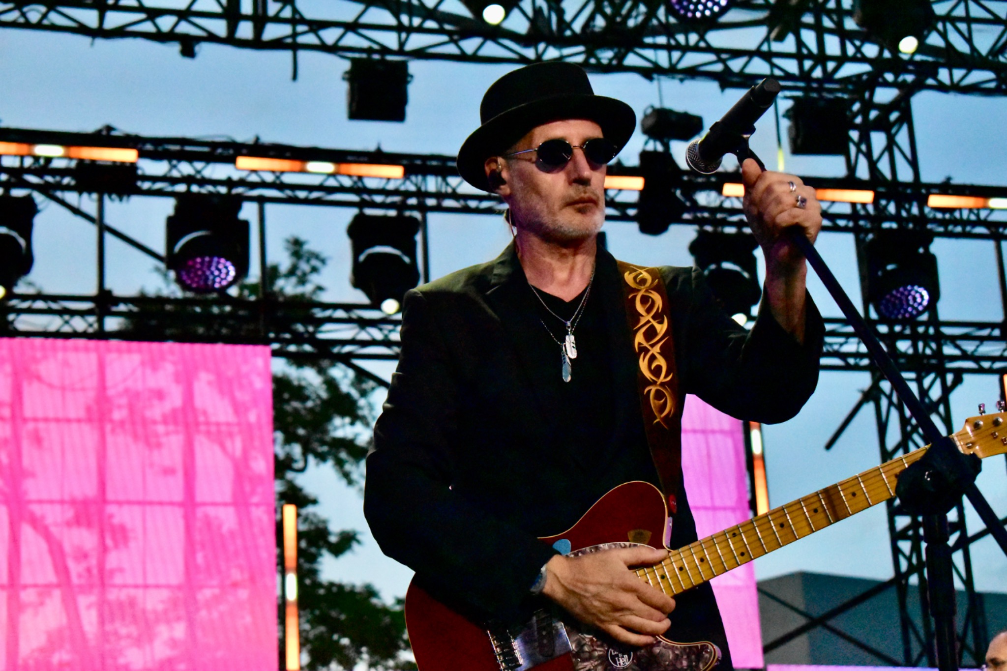 Mężczyzna w kapeluszu i okularach gra na gitarze podczas koncertu, w tle konstrukcja sceniczna i oświetlenie. Wokalista trzyma mikrofon w dłoni.