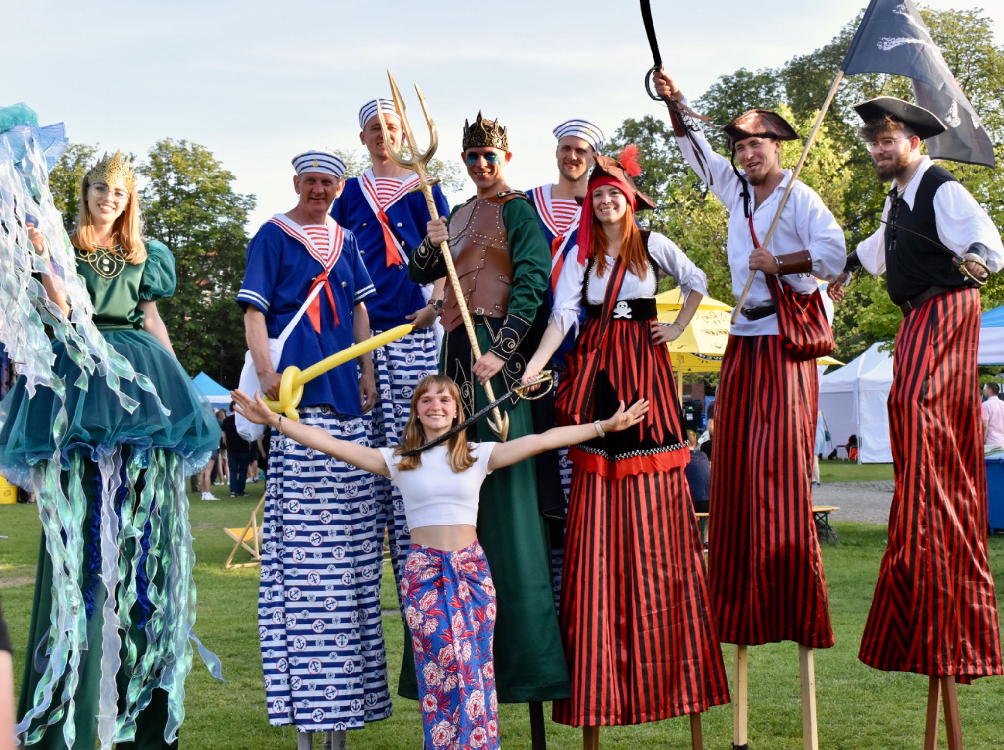 Grupa osób w kostiumach: żeglarzy, piratów, meduzy i Neptuna, pozuje na łące. Niektórzy na szczudłach. Jeden trzyma flagę piracką, inny trójząb. Uśmiechnięci, w plenerze.