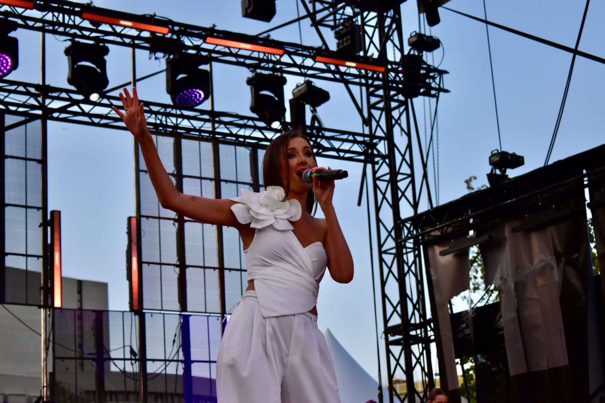 Piosenkarka w białym stroju śpiewa na scenie podczas koncertu plenerowego. W tle konstrukcja sceniczna i oświetlenie.