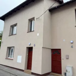 Elewacja bliźniaka w beżowym kolorze z czerwonymi drzwiami wejściowymi i numerami 4 i 6, widok z zewnątrz na tle pochmurnego nieba i zieleni.