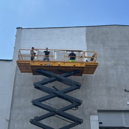 Pw Bartosz Olszewski - Trzech malarzy na podnośniku maluje elewację budynku na szaro, widoczna część pomalowana i niepomalowana, jasne niebo, budynek z numerem 6B.