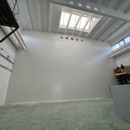 Pw Bartosz Olszewski - Wnętrze hali z białymi ścianami z płyt, oświetlone przez okna dachowe i boczne. Podnośnik koszowy z pracownikami przy ścianie. Widoczna surowa podłoga.