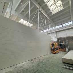 Pw Bartosz Olszewski - Montaż płyt warstwowych na ścianie hali przemysłowej. Pracownicy na podnośnikach koszowych. Widoczne płyty do montażu i elementy konstrukcji dachu.