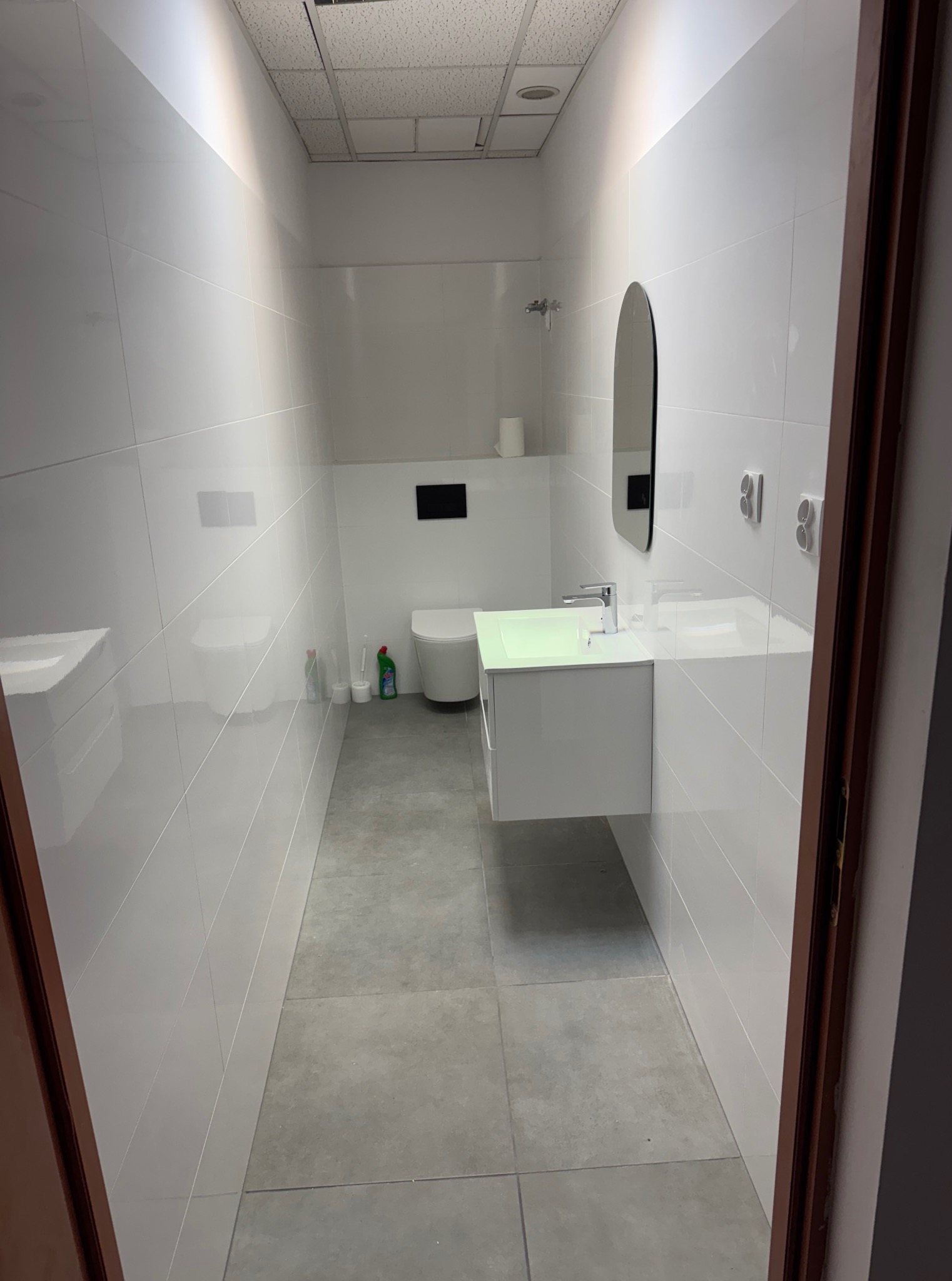 Nowoczesna łazienka po remoncie z białymi płytkami, owalnym lustrem i podwieszaną szafką z umywalką. Widok zza uchylonych, ciemnych drzwi.