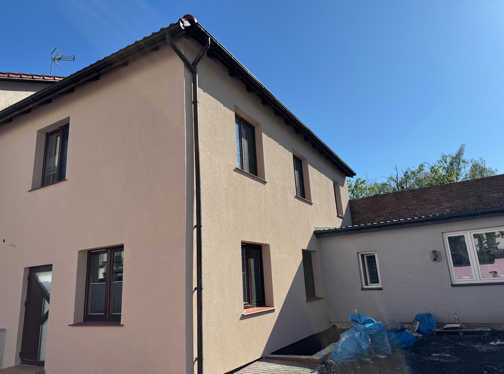 Ocieplony dom jednorodzinny w Inowrocławiu z nową elewacją w odcieniach beżu, widoczny z boku, z ciemnymi oknami i dachem. W tle błękitne niebo i zieleń drzew.