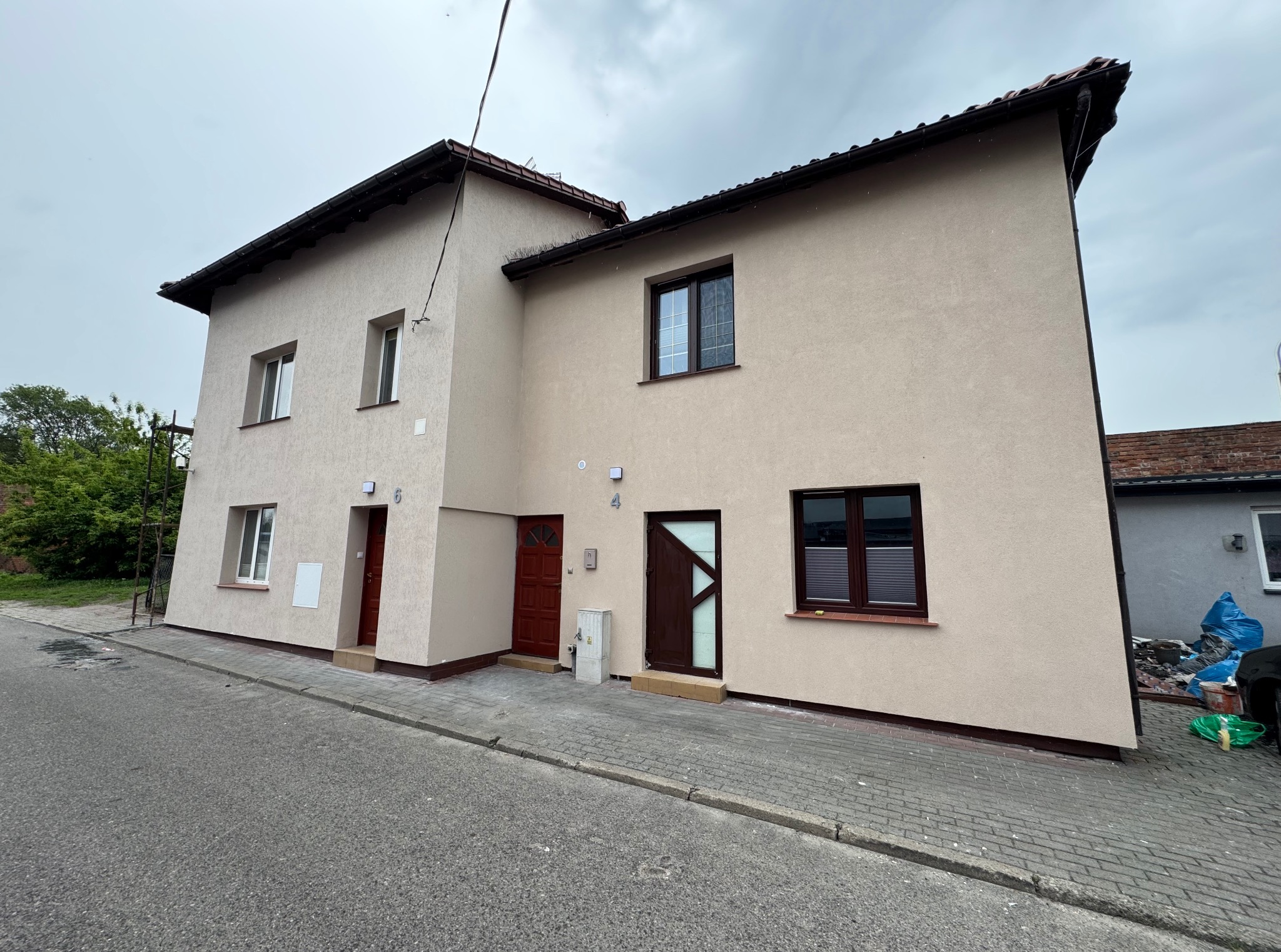 Dwukondygnacyjny budynek bliźniaczy po remoncie elewacji w beżowej tonacji, z brązowymi oknami i drzwiami. Widok z poziomu ulicy, pochmurne niebo.