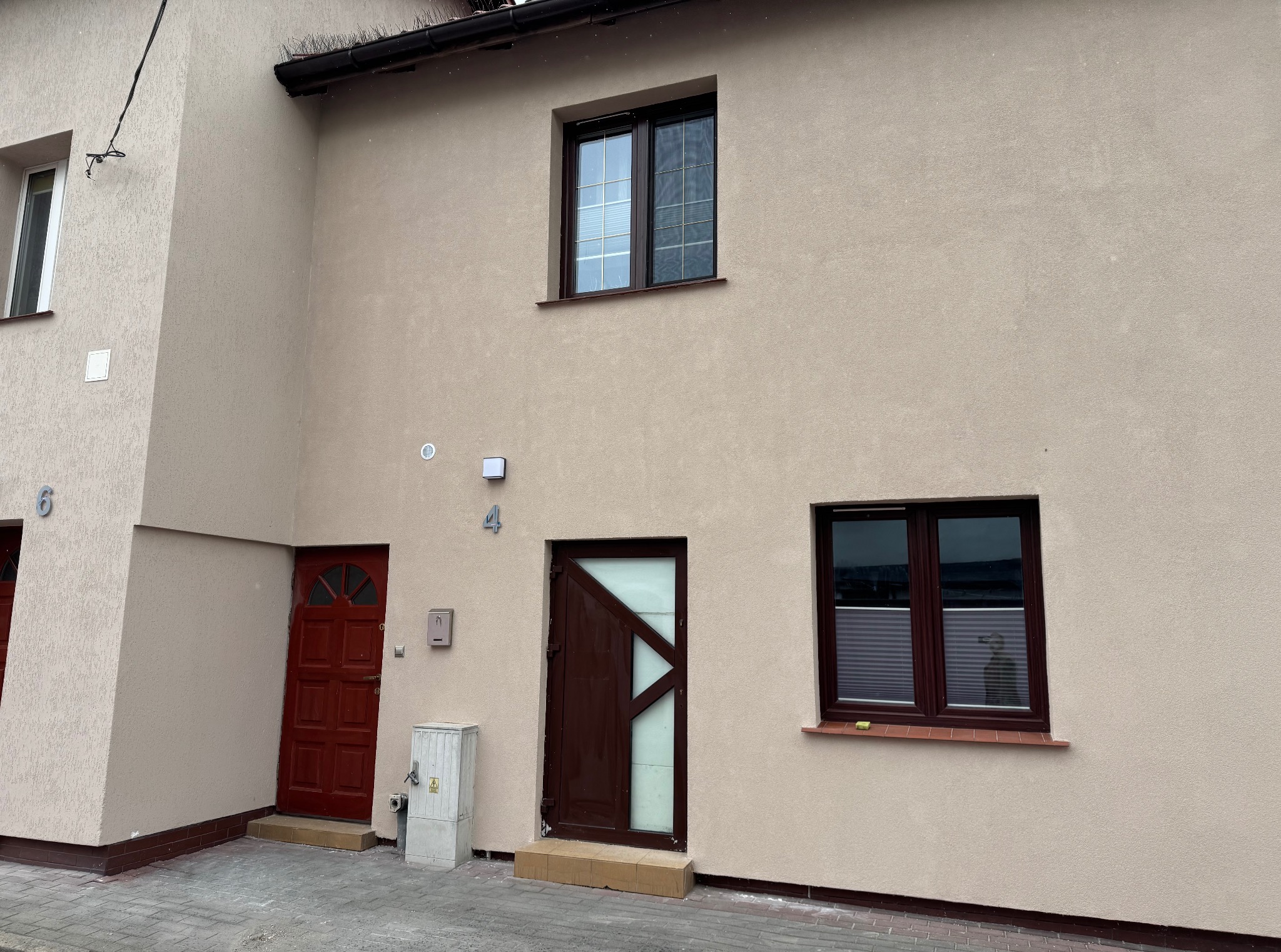 Elewacja budynku szeregowego z beżowym tynkiem, ciemnymi oknami i drzwiami. Widoczne numery domów 4 i 6. Na dachu system zabezpieczeń przed ptakami.