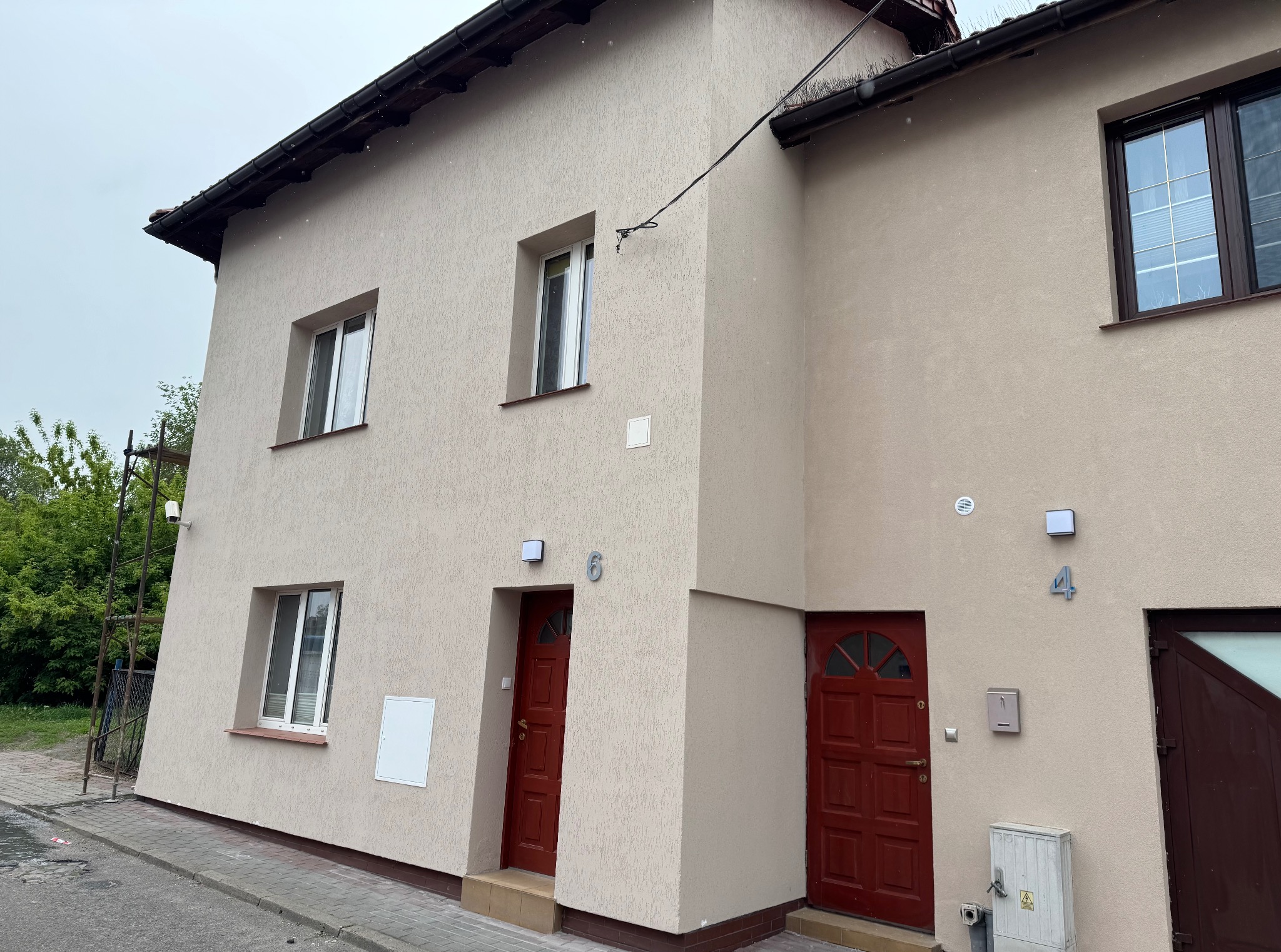 Elewacja bliźniaka w beżowym kolorze z czerwonymi drzwiami wejściowymi i numerami 4 i 6, widok z zewnątrz na tle pochmurnego nieba i zieleni.