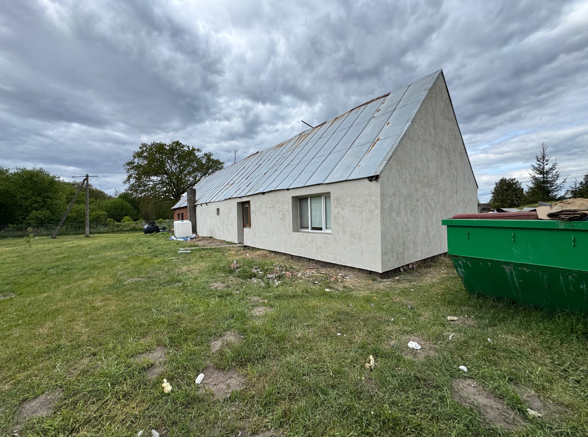 Elewacja domu po remoncie, nowa, jasna, z metalowym dachem. Na trawniku stoi zielony kontener na odpady. Pochmurne niebo w tle.