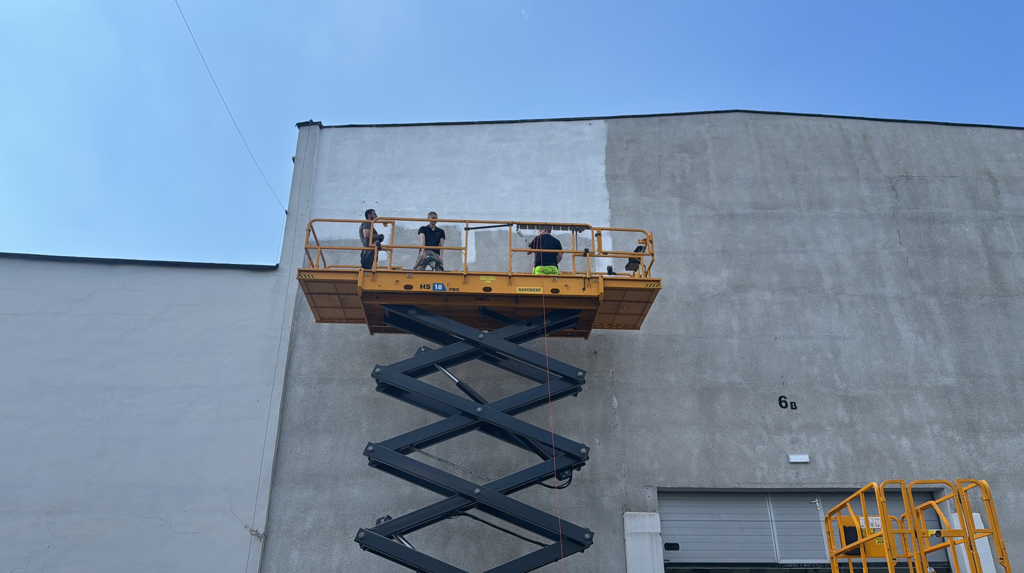Trzech malarzy na podnośniku maluje elewację budynku na szaro, widoczna część pomalowana i niepomalowana, jasne niebo, budynek z numerem 6B.