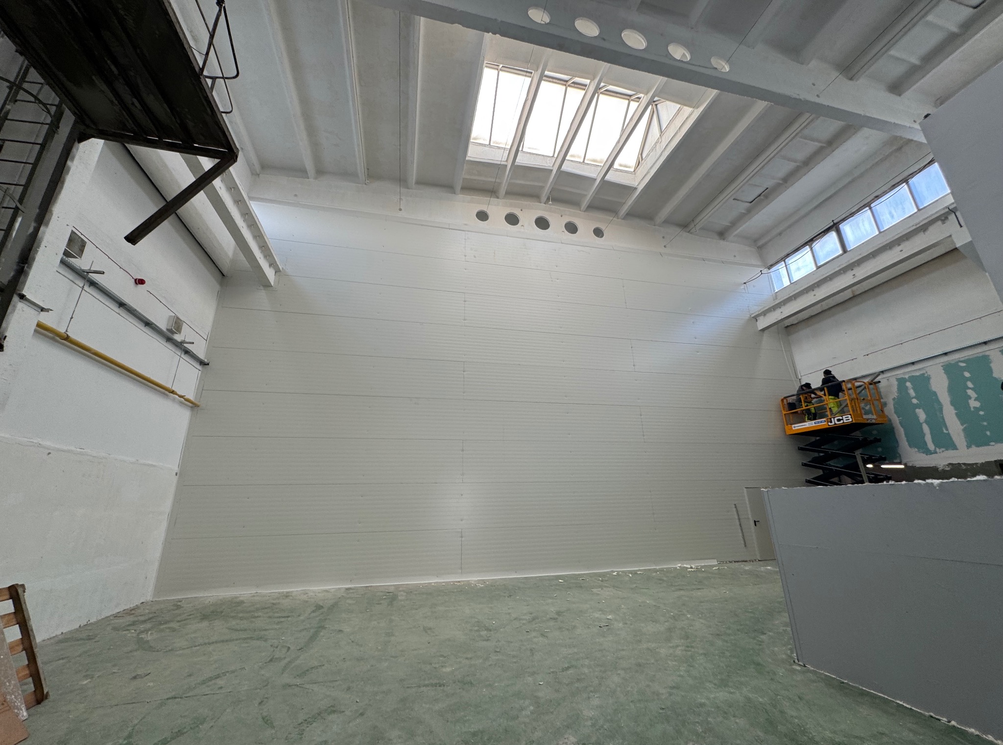 Wnętrze hali z białymi ścianami z płyt, oświetlone przez okna dachowe i boczne. Podnośnik koszowy z pracownikami przy ścianie. Widoczna surowa podłoga.