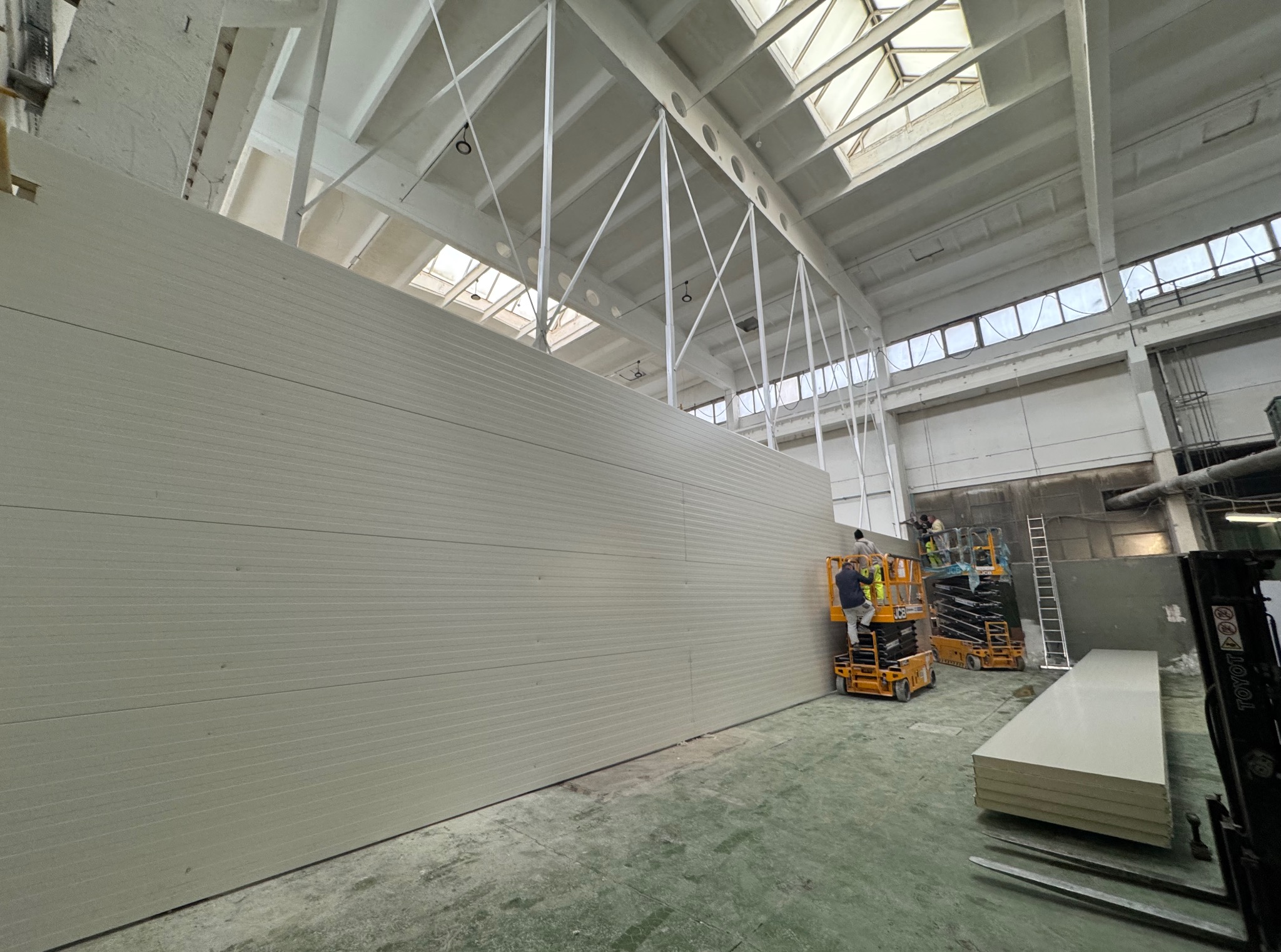 Montaż płyt warstwowych na ścianie hali przemysłowej. Pracownicy na podnośnikach koszowych. Widoczne płyty do montażu i elementy konstrukcji dachu.