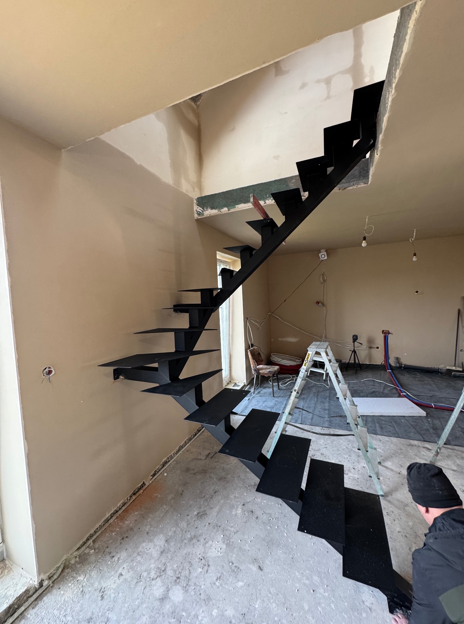 Czarne, ażurowe schody zamontowane w surowym wnętrzu. Widoczne ślady prac remontowych, drabina i instalacje. Ściany w odcieniach beżu, betonowa podłoga.