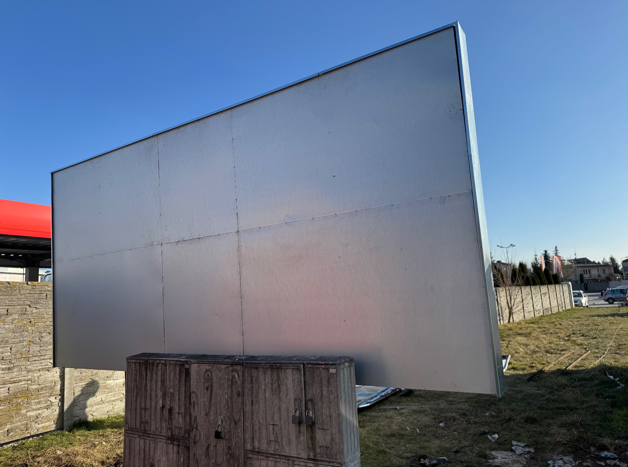 Billboard z metalową konstrukcją na tle błękitnego nieba i zabudowań. Widoczne elementy mocujące i skrzynka elektryczna u podstawy.
