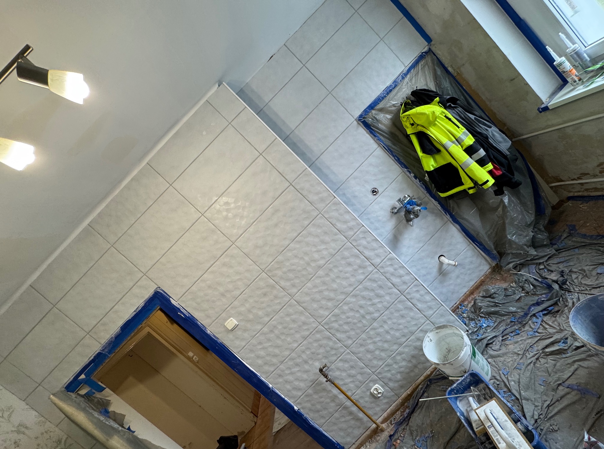 Świeżo wykafelkowana łazienka z jasnymi, kwadratowymi płytkami. Widoczne narzędzia glazurnicze, wiadro i odzież robocza. Kąt ujęcia z góry podkreśla ukończony fragment ściany.