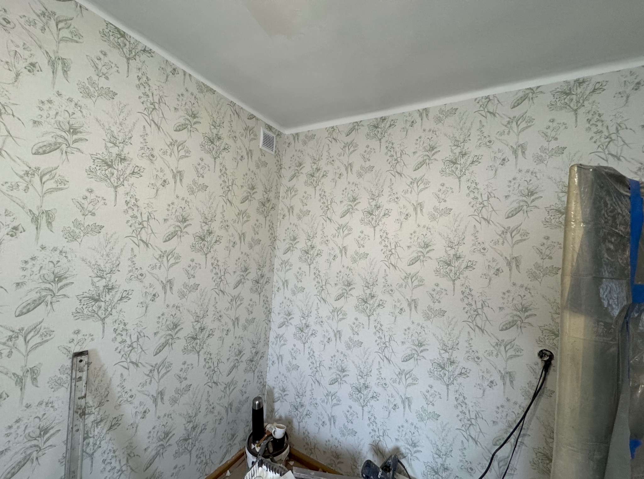 Pokój po tapetowaniu w delikatny wzór roślinny. Widoczne narzędzia malarskie i zabezpieczone elementy. Świeżo położona tapeta z motywem kwiatowym.