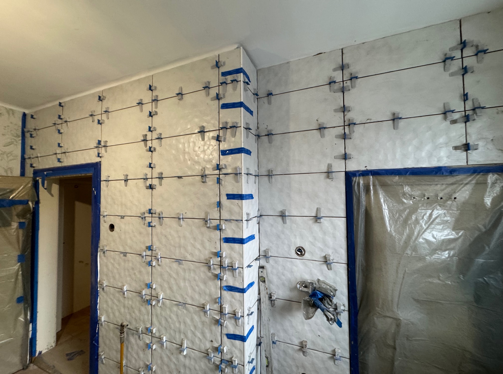 Ściana w trakcie układania białych, błyszczących płytek z użyciem klipsów poziomujących i taśmy malarskiej, widoczne zabezpieczone folią drzwi.