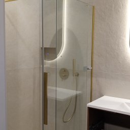 Daniel Zięba - Nowoczesna kabina prysznicowa z elementami w kolorze złota, oświetleniem LED i deszczownicą. Umywalka z szafką w tle. Jasne płytki na ścianach i podłodze.