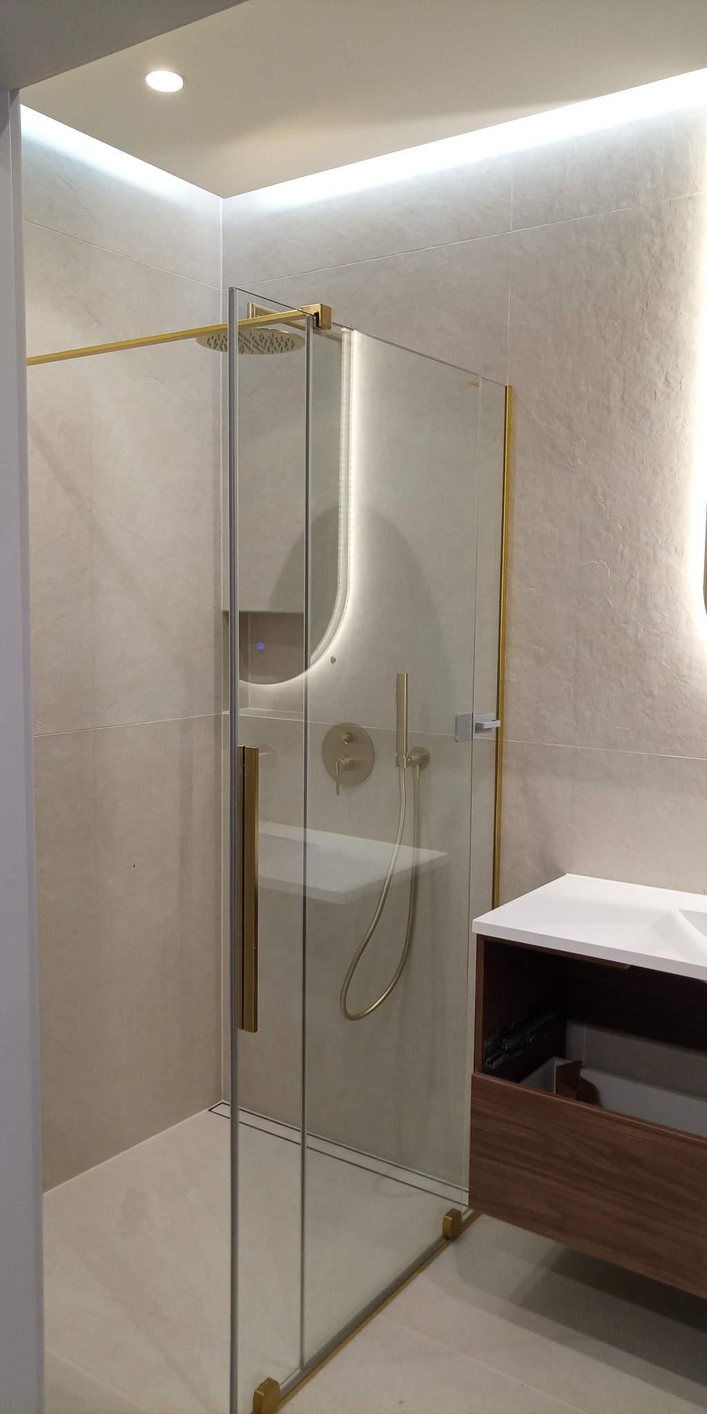 Nowoczesna kabina prysznicowa z elementami w kolorze złota, oświetleniem LED i deszczownicą. Umywalka z szafką w tle. Jasne płytki na ścianach i podłodze.