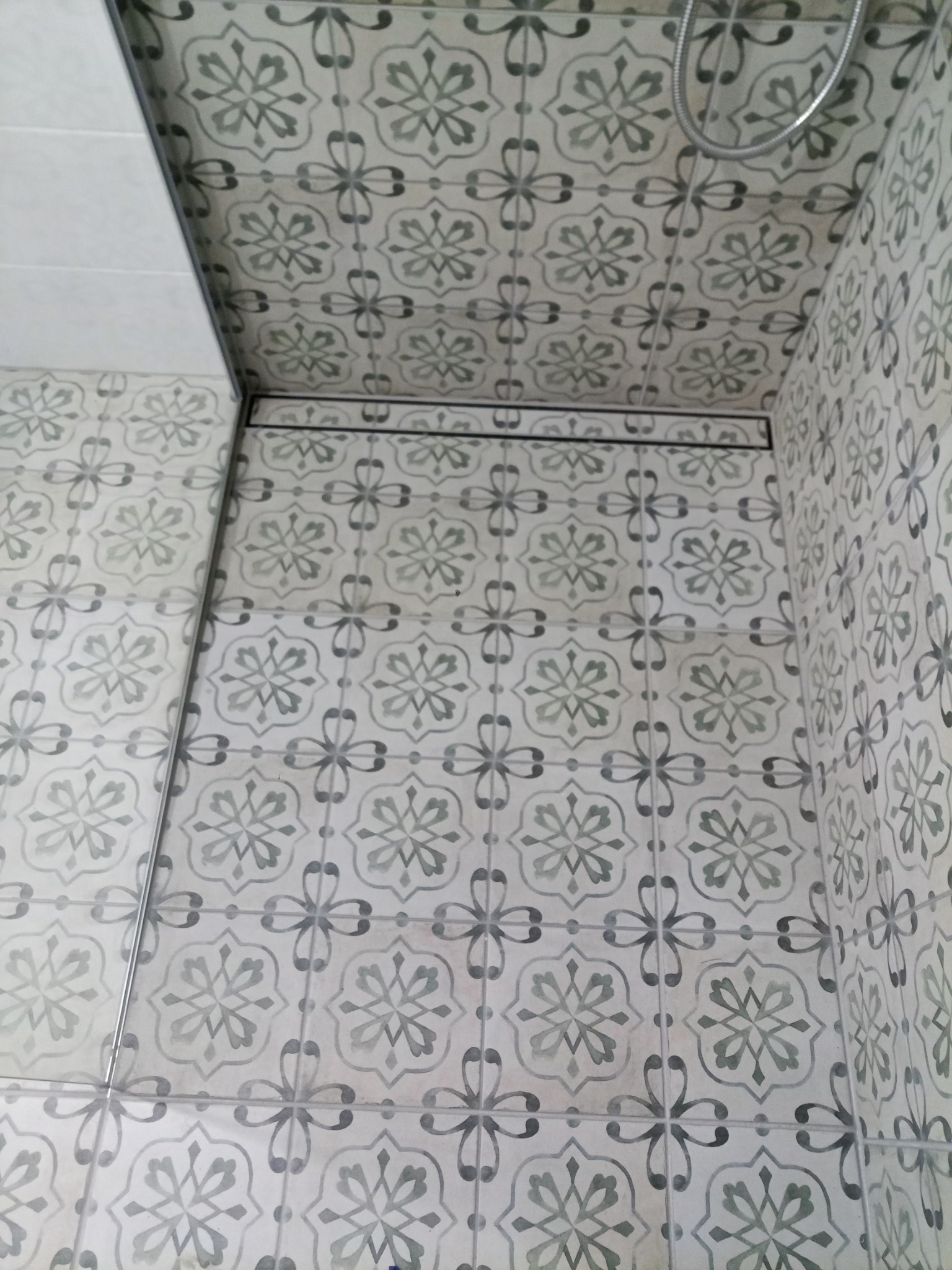 Wykończona kabina prysznicowa z dekoracyjnymi, wzorzystymi płytkami w odcieniach szarości i bieli, widoczny odpływ liniowy i słuchawka prysznicowa.