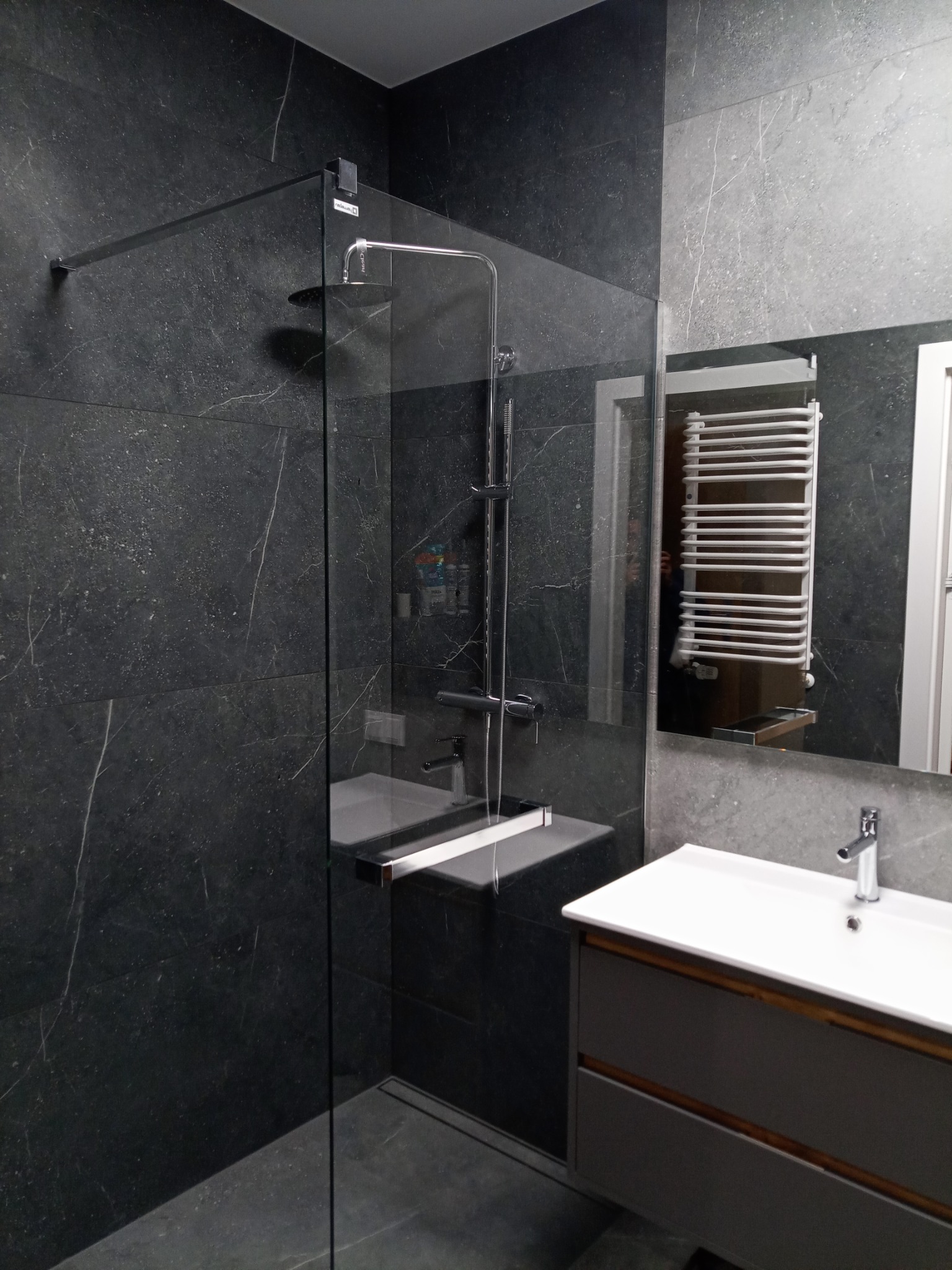 Nowoczesna łazienka z kabiną prysznicową typu walk-in, wykończona ciemnymi płytkami imitującymi marmur. Widoczna umywalka z szafką i grzejnik drabinkowy w lustrze.