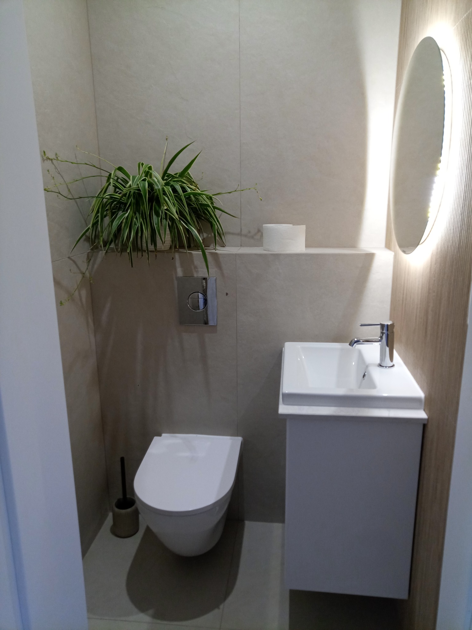Nowoczesna łazienka z podwieszaną toaletą, umywalką na szafce i okrągłym lustrem z podświetleniem LED. Na ścianie roślina doniczkowa.
