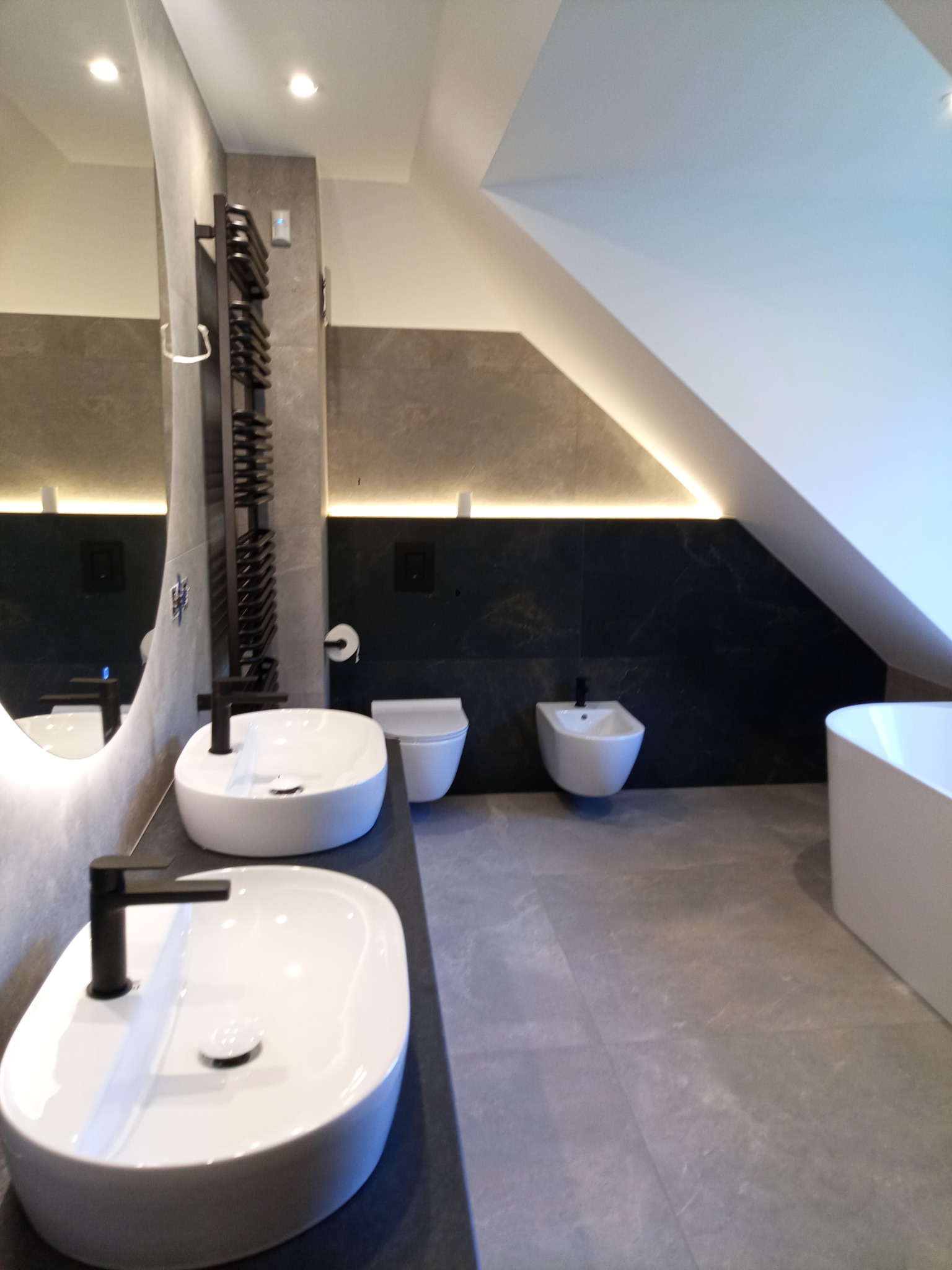 Nowoczesna łazienka na poddaszu z dwoma umywalkami, wiszącą toaletą i bidetem. Szare płytki, czarne akcenty i oświetlenie LED tworzą elegancki klimat.